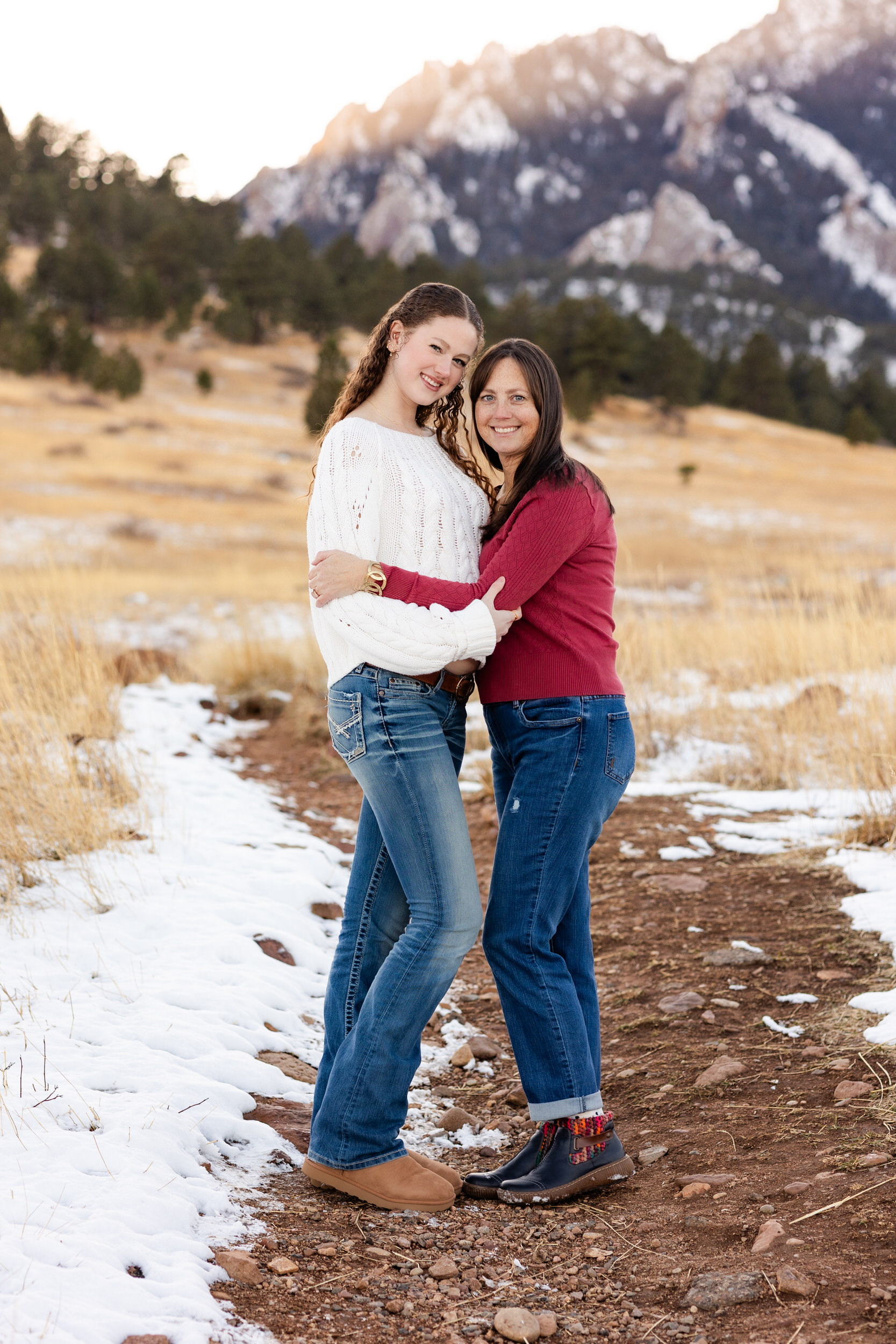 A mom and her adult daughter hug each other and smile at the camera.