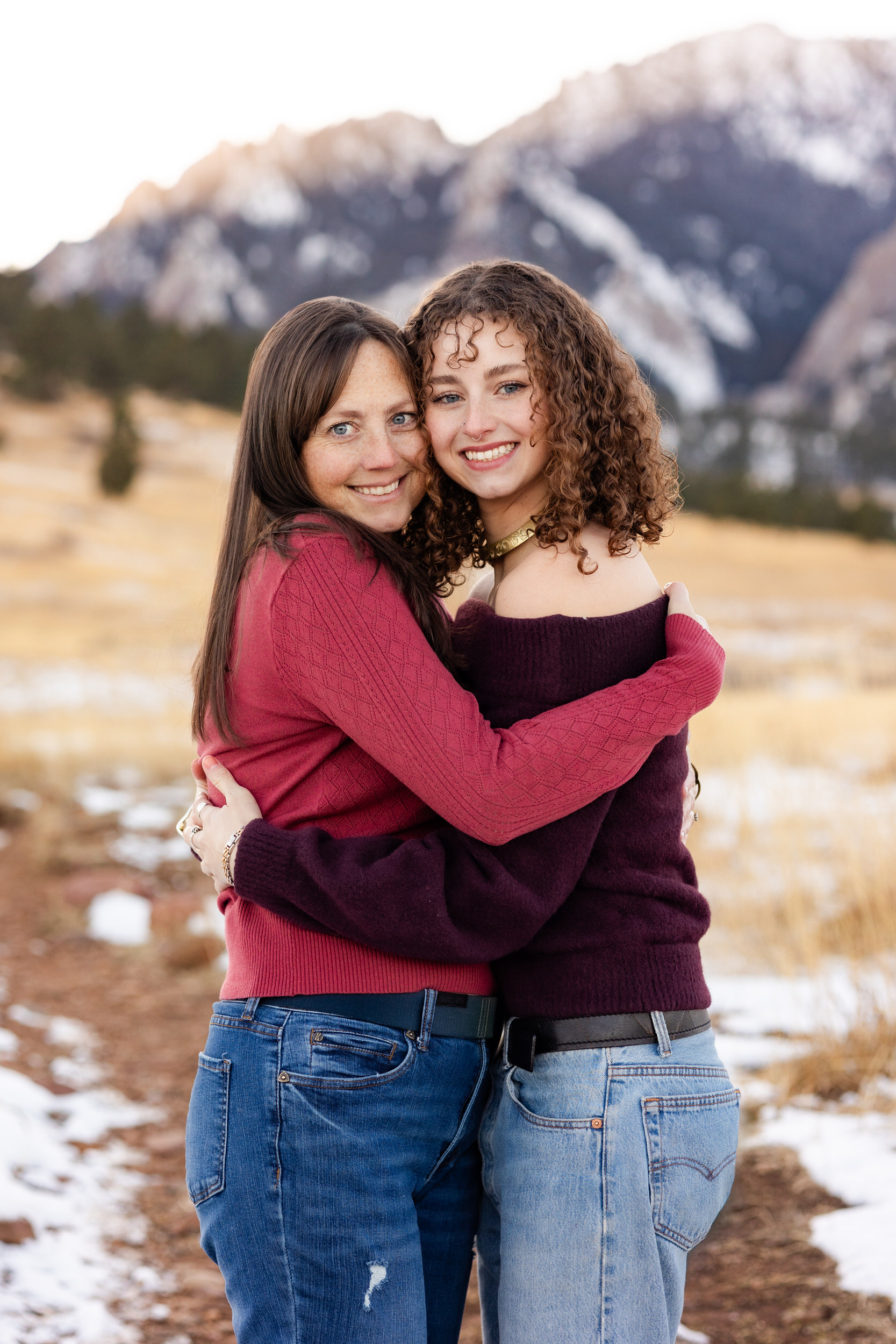 A mom and her adult daughter hug each other and smile at the camera.