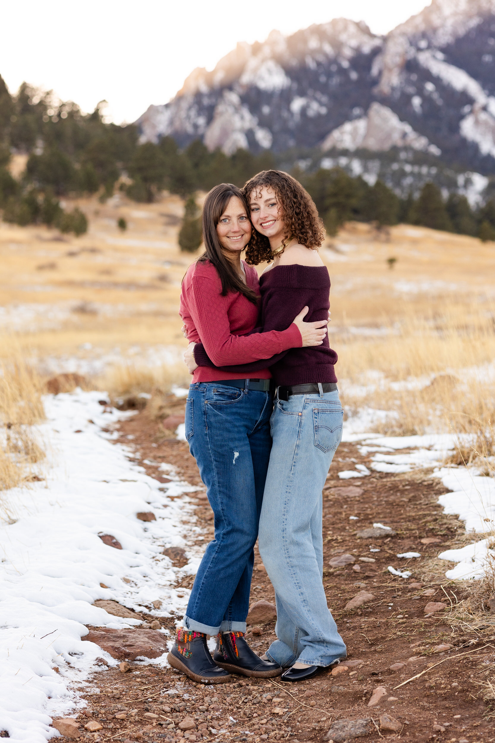 A mom and her adult daughter hug each other and smile at the camera.