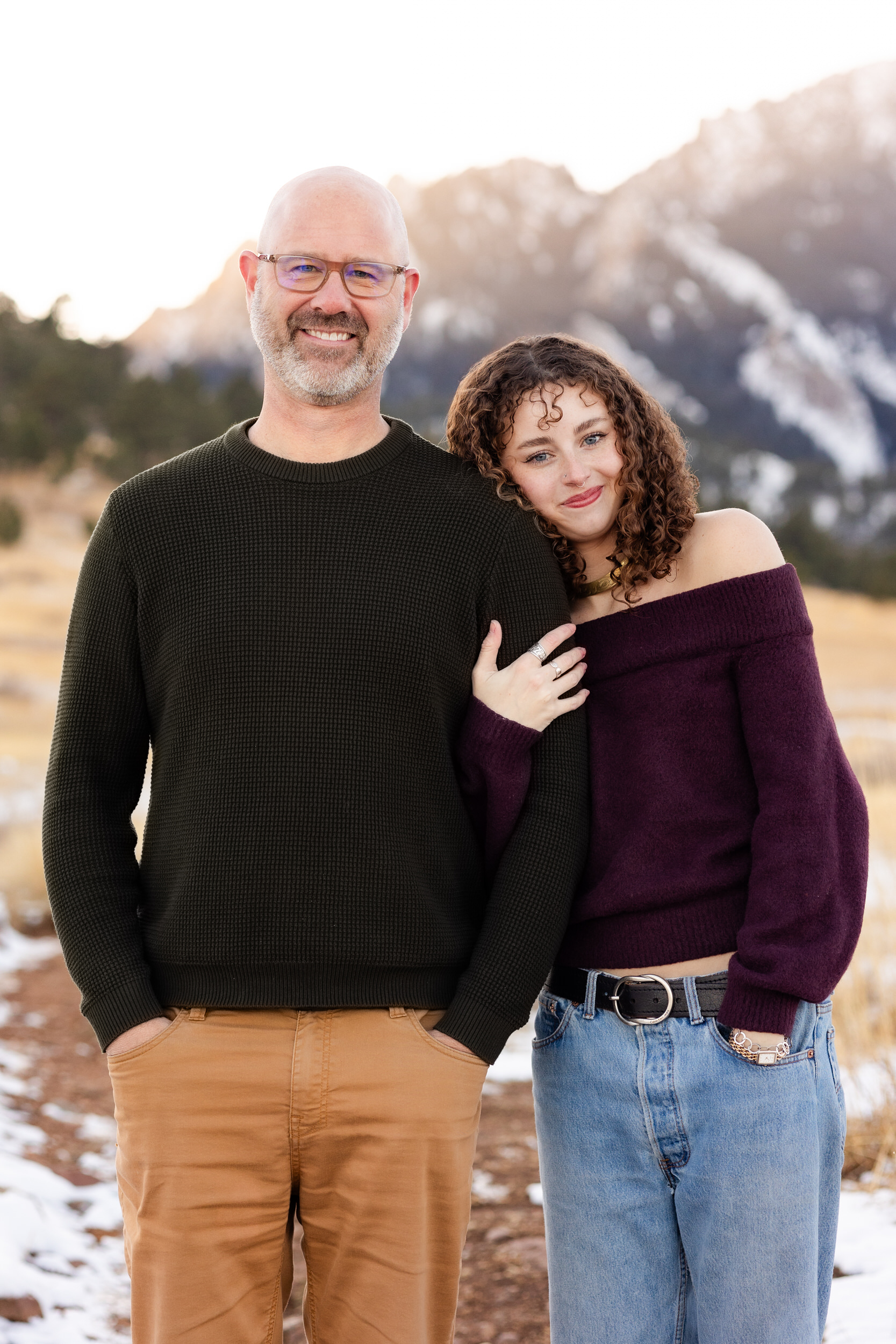 A young woman stands arm-in-arm with her dad and leans her head on his shoulder as they both smile at the camera.