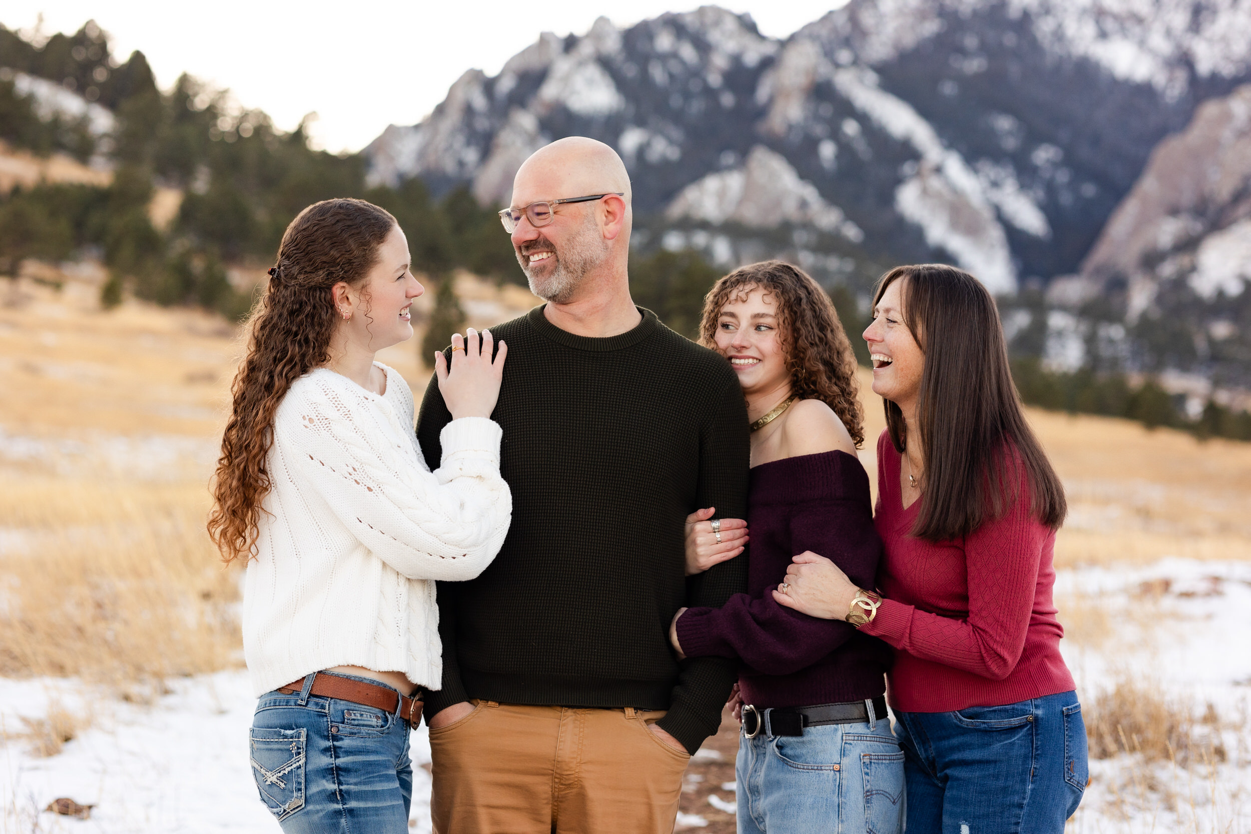 A mom and dad stand arm-in-arm with their two young adult daughters and they all smile at each other.
