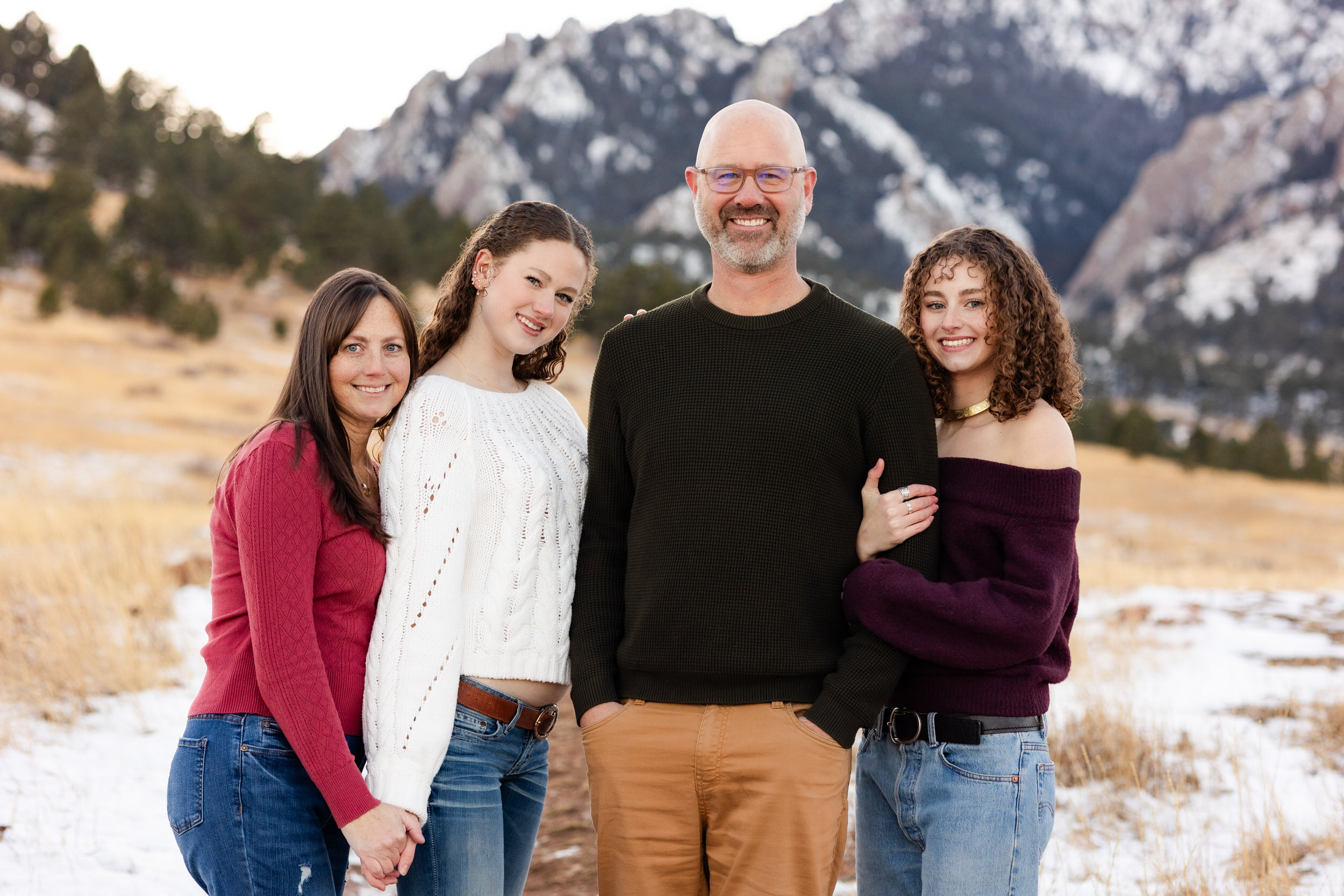 A mom and dad stand arm-in-arm with their two young adult daughters and they all smile at the camera.