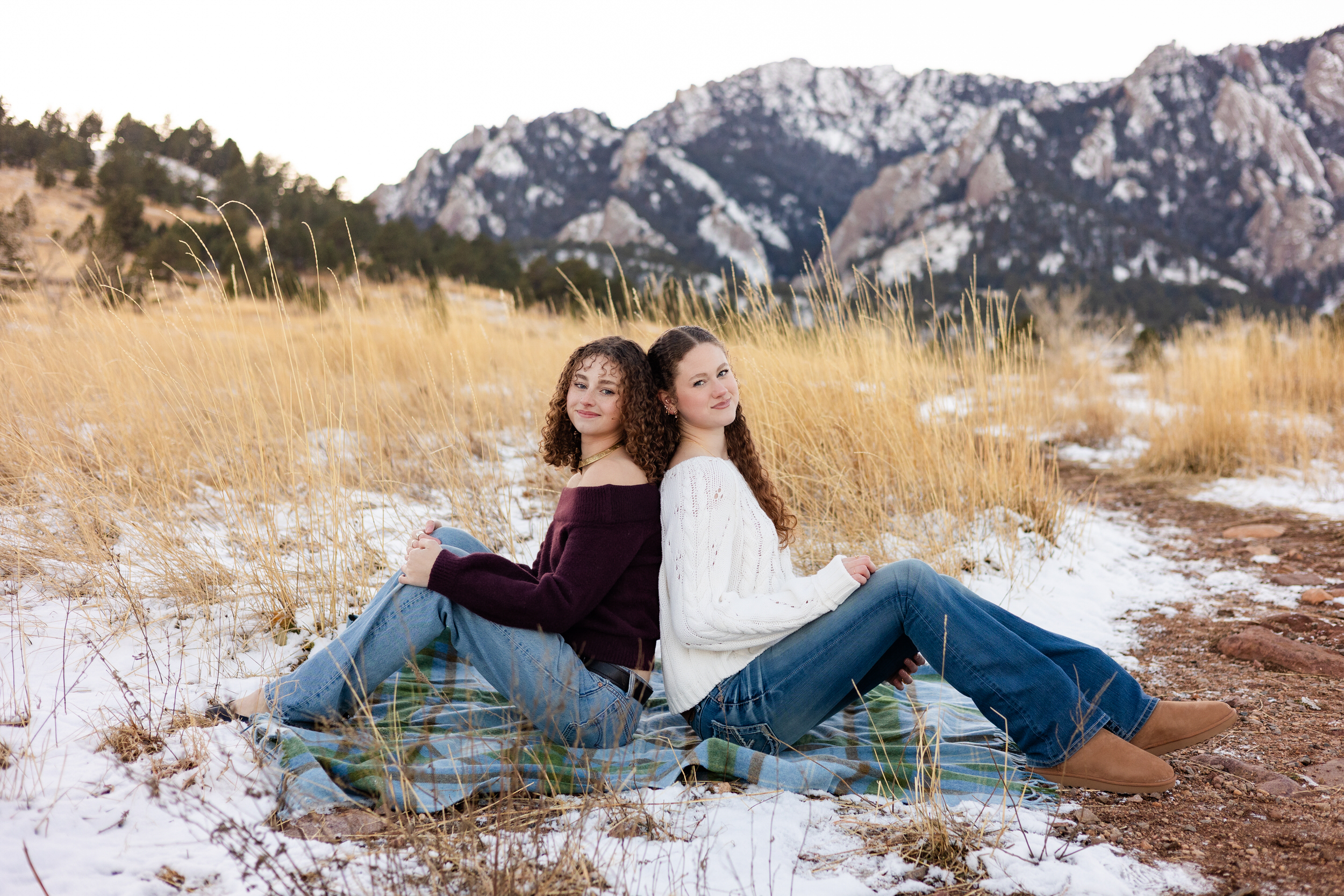 Two young women sit on a blanket back to back and smile at the camera.