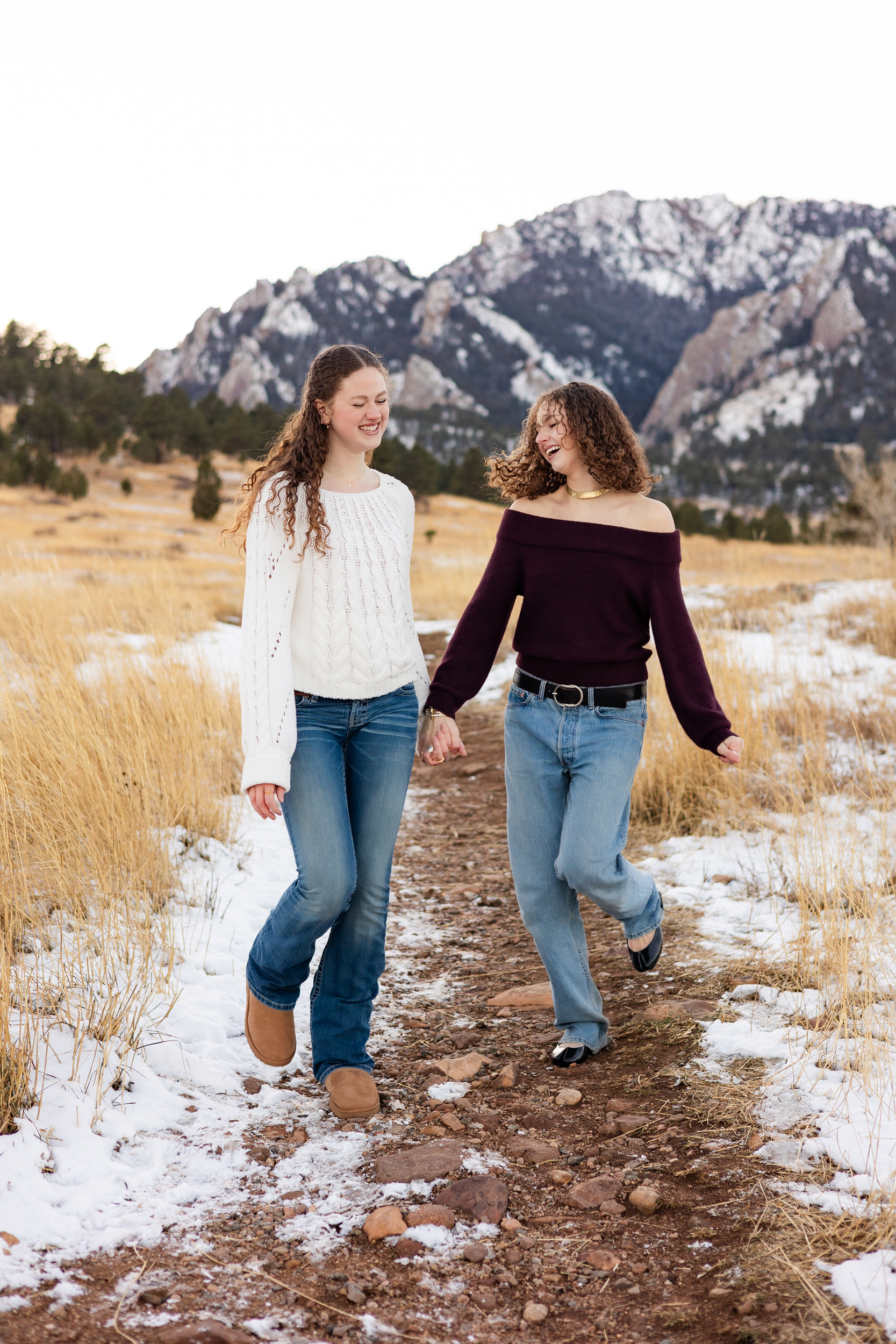 Two young women hold hands and run down a dirt path as they laugh together.