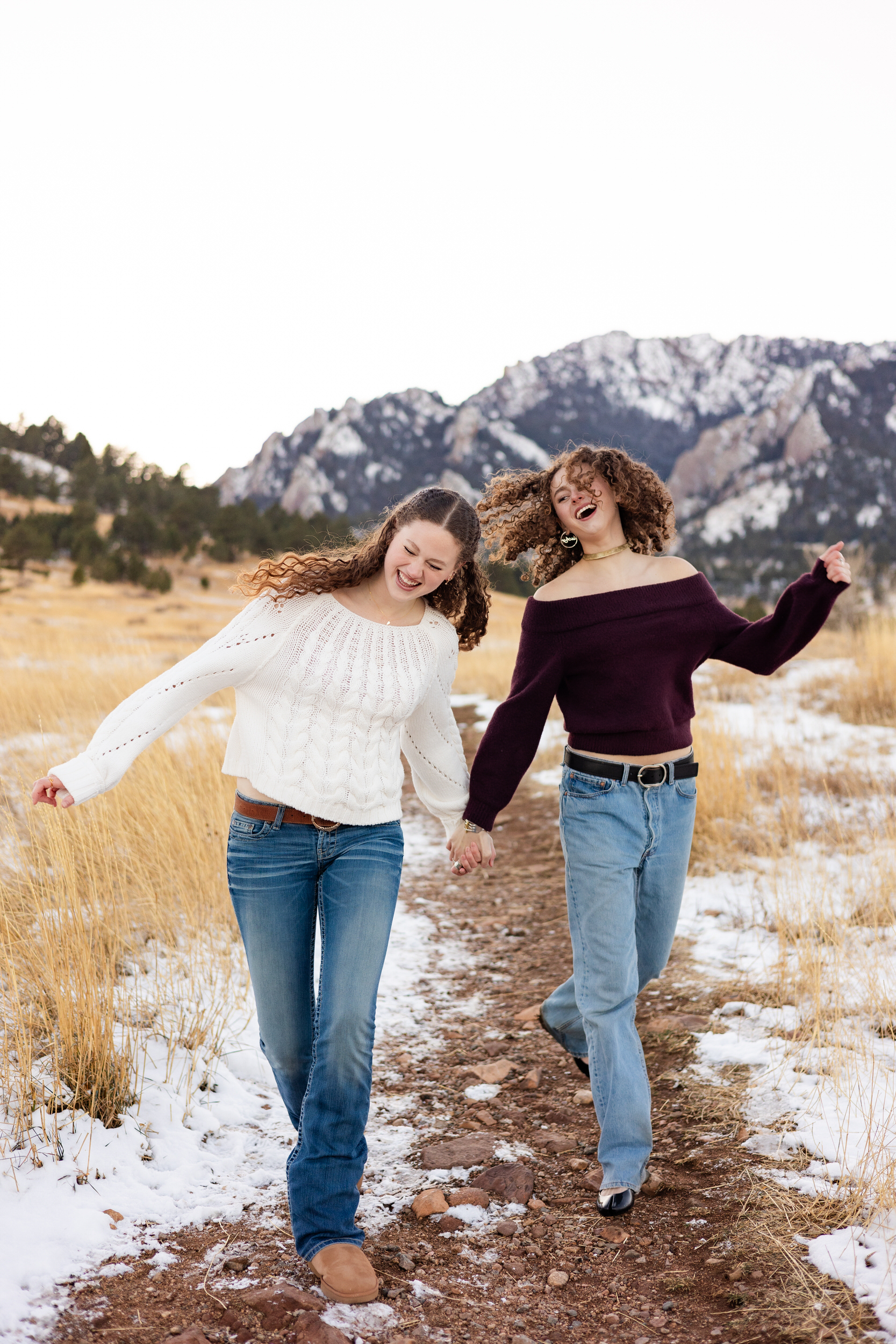 Two young women hold hands as they skip down a dirt path and laugh together.