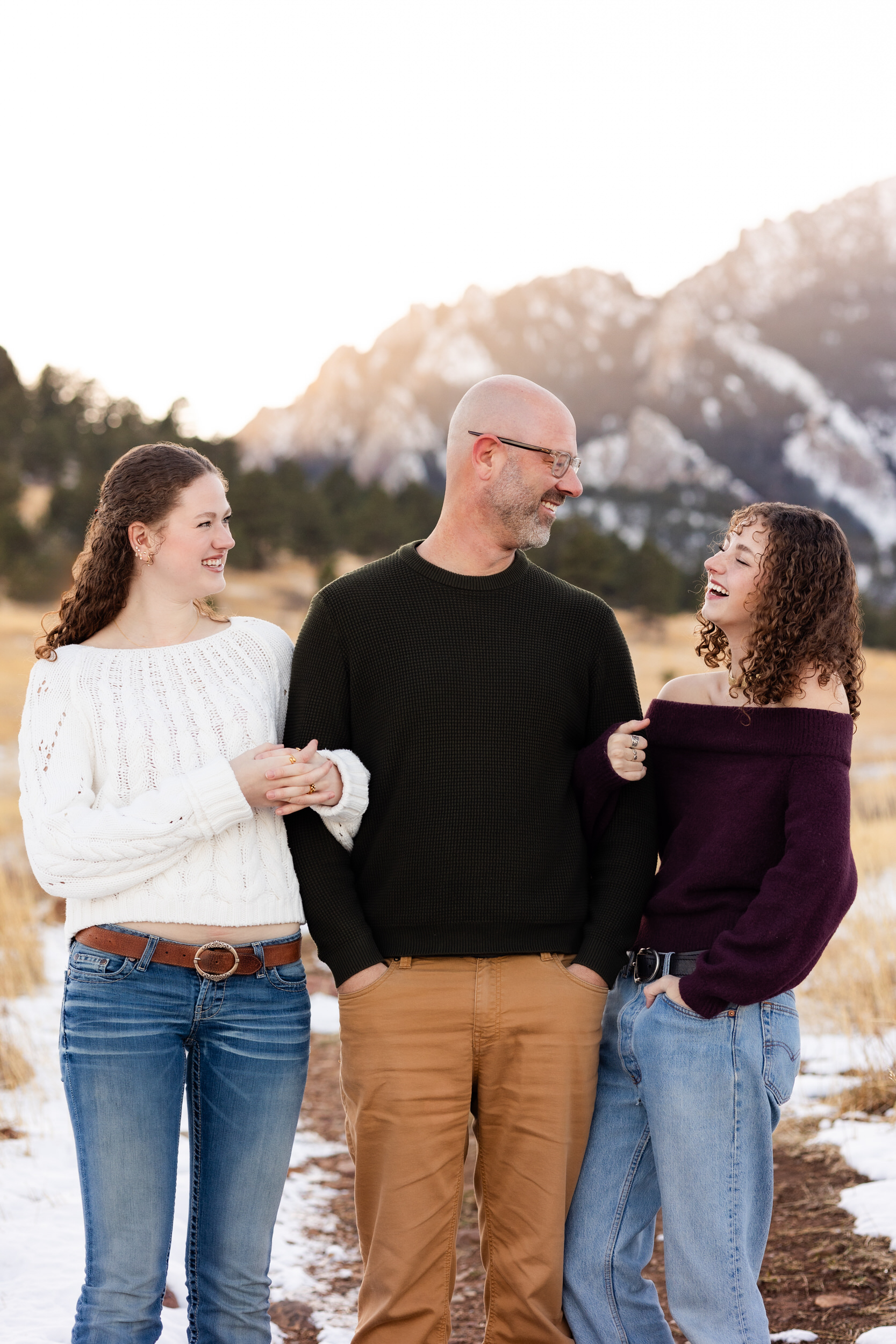 Two sisters stand arm-in-arm with their dad as they laugh together.