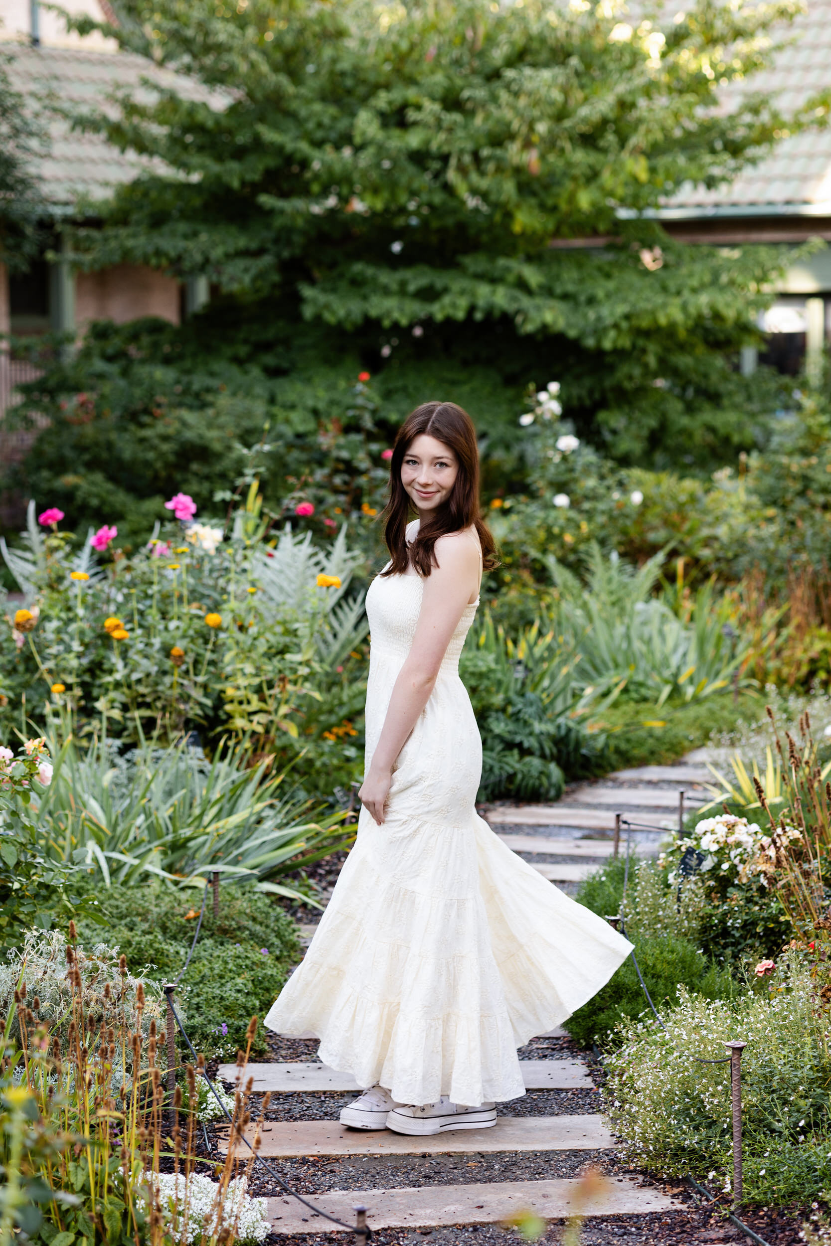 A young woman in a long white dress swishes her skirt and looks at the camera while standing on a path at the Denver Botanic Gardens.