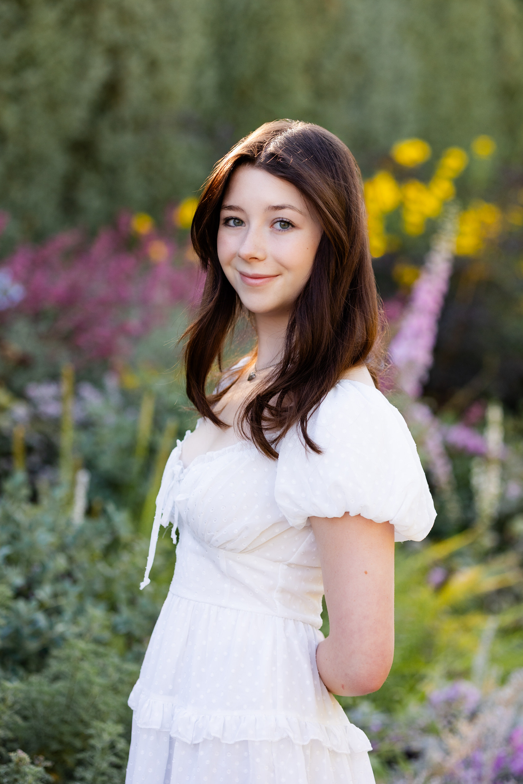 A young woman in a white dress stands in front of flowers at the Denver Botanic Gardens and smiles at the camera.