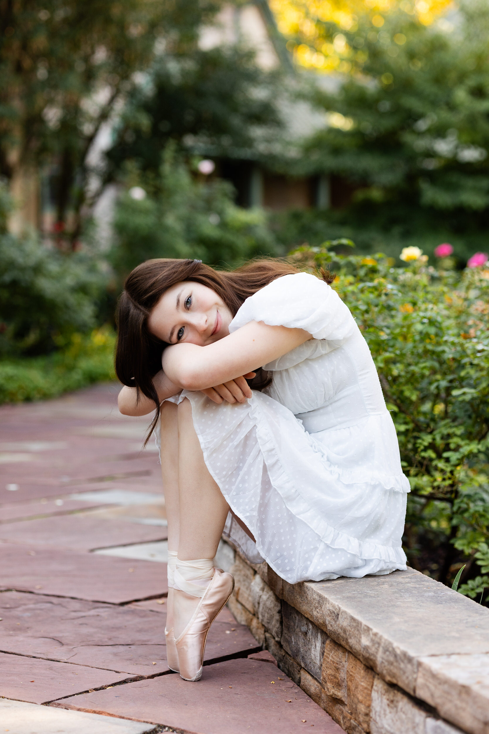 A young woman sits on a ledge in a garden and rests her head on her knees with her feet en pointe as she smiles at the camera.