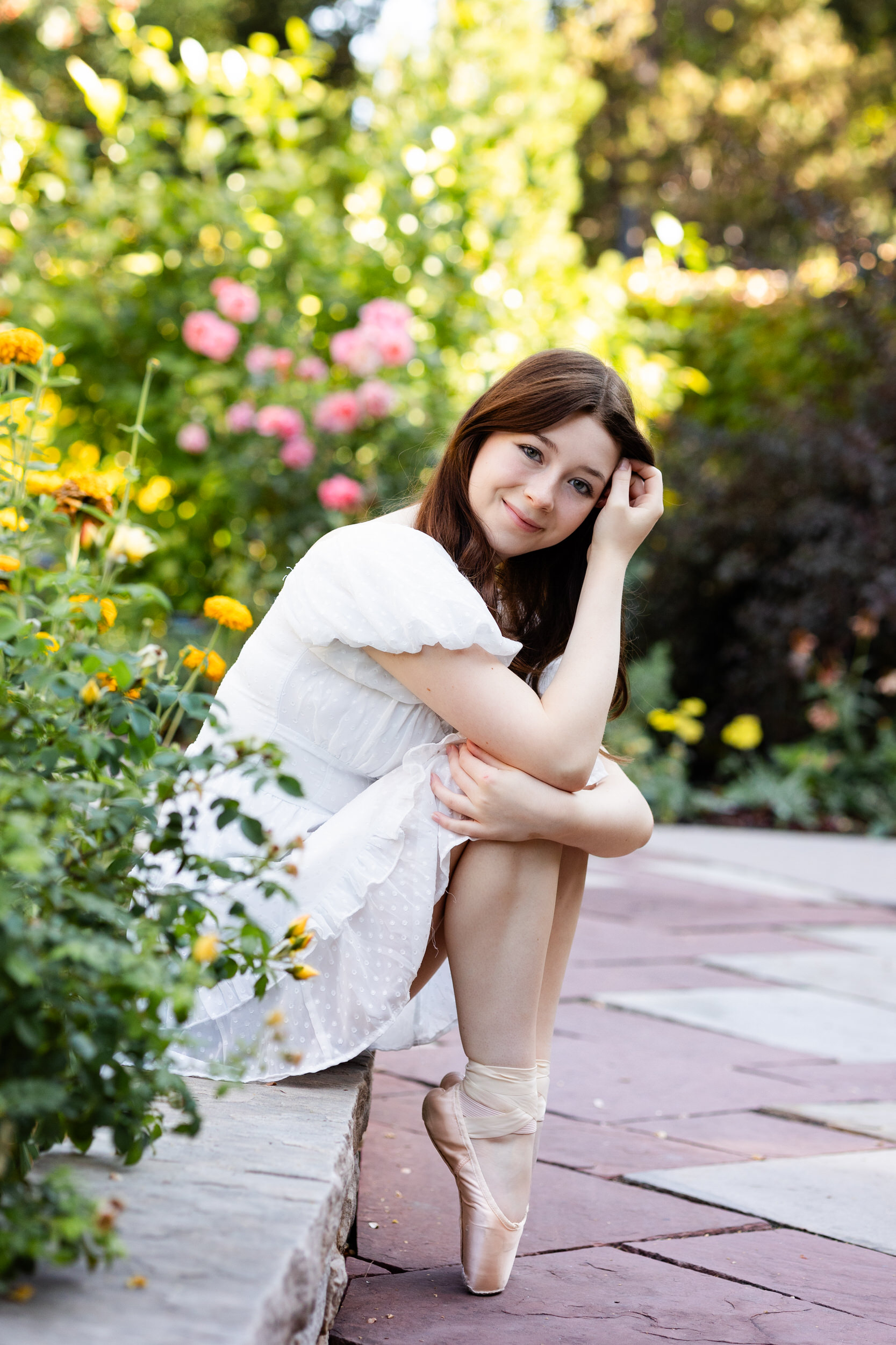 A young woman sits on a ledge in a garden with her feet en pointe and runs one hand through her hair as she smiles at the camera.