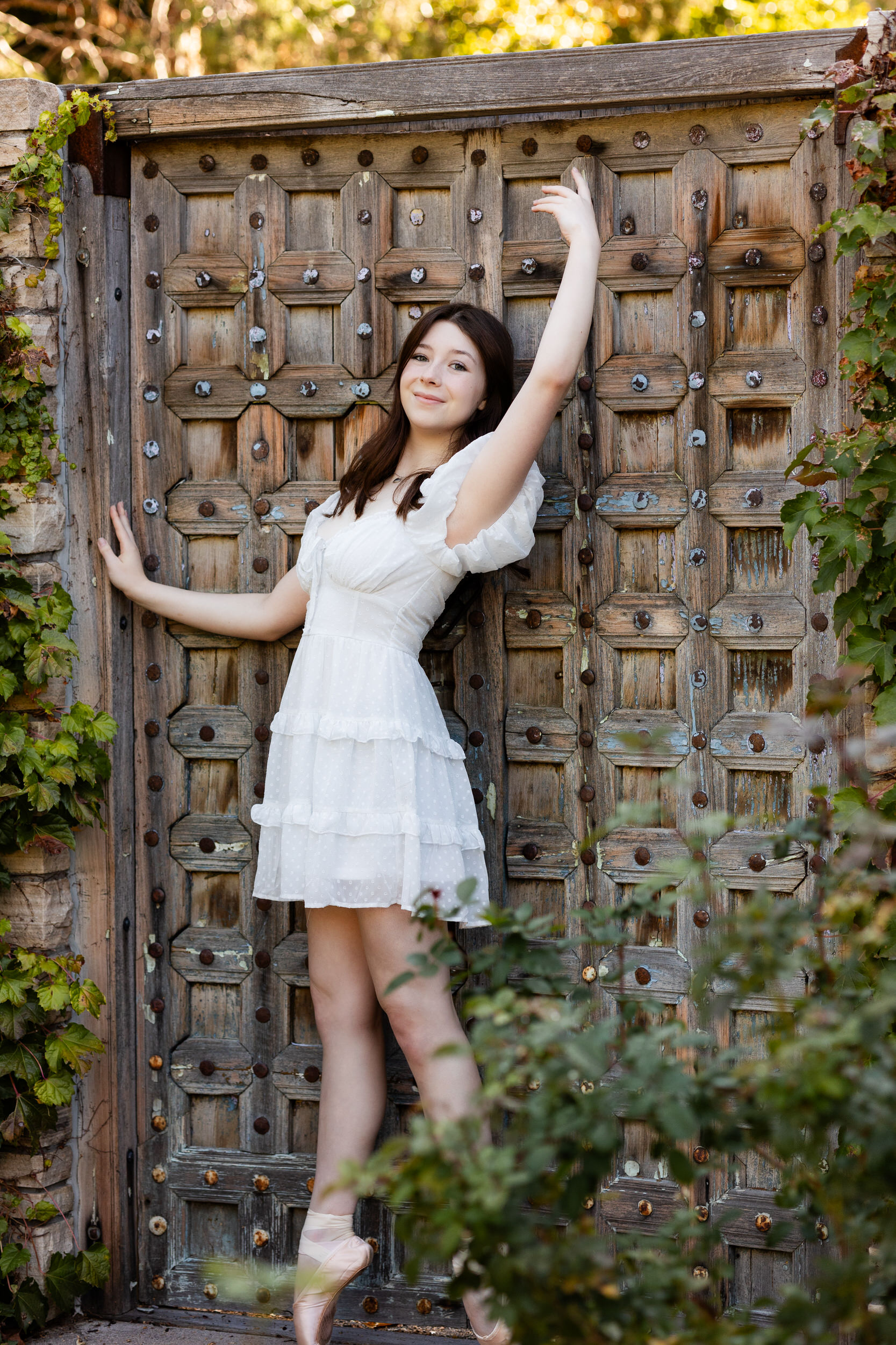 A young woman in a white dress stands en pointe in front of a garden door.