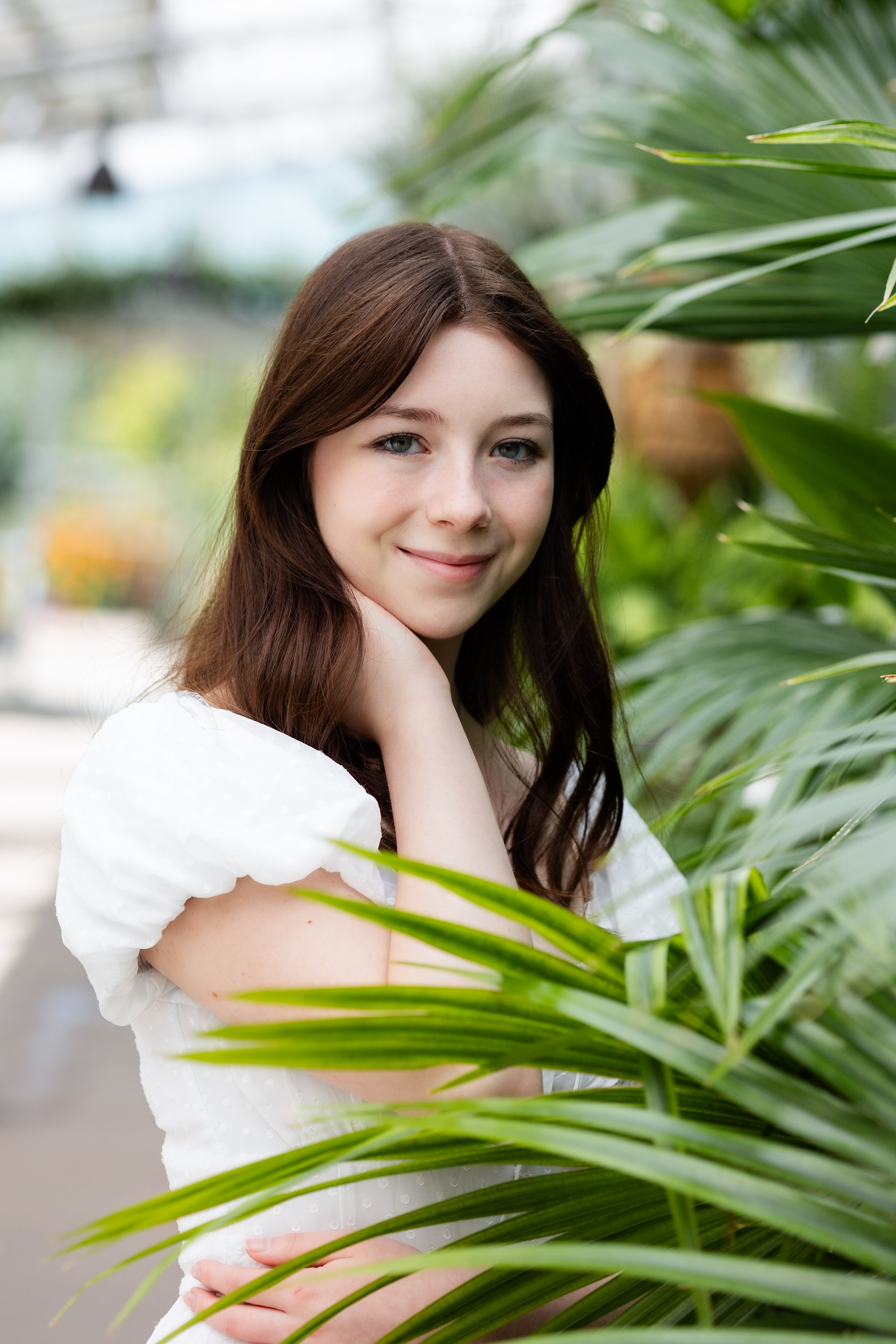 A young woman standing in a greenhouse runs one hand through her hair as she smiles at the camera.