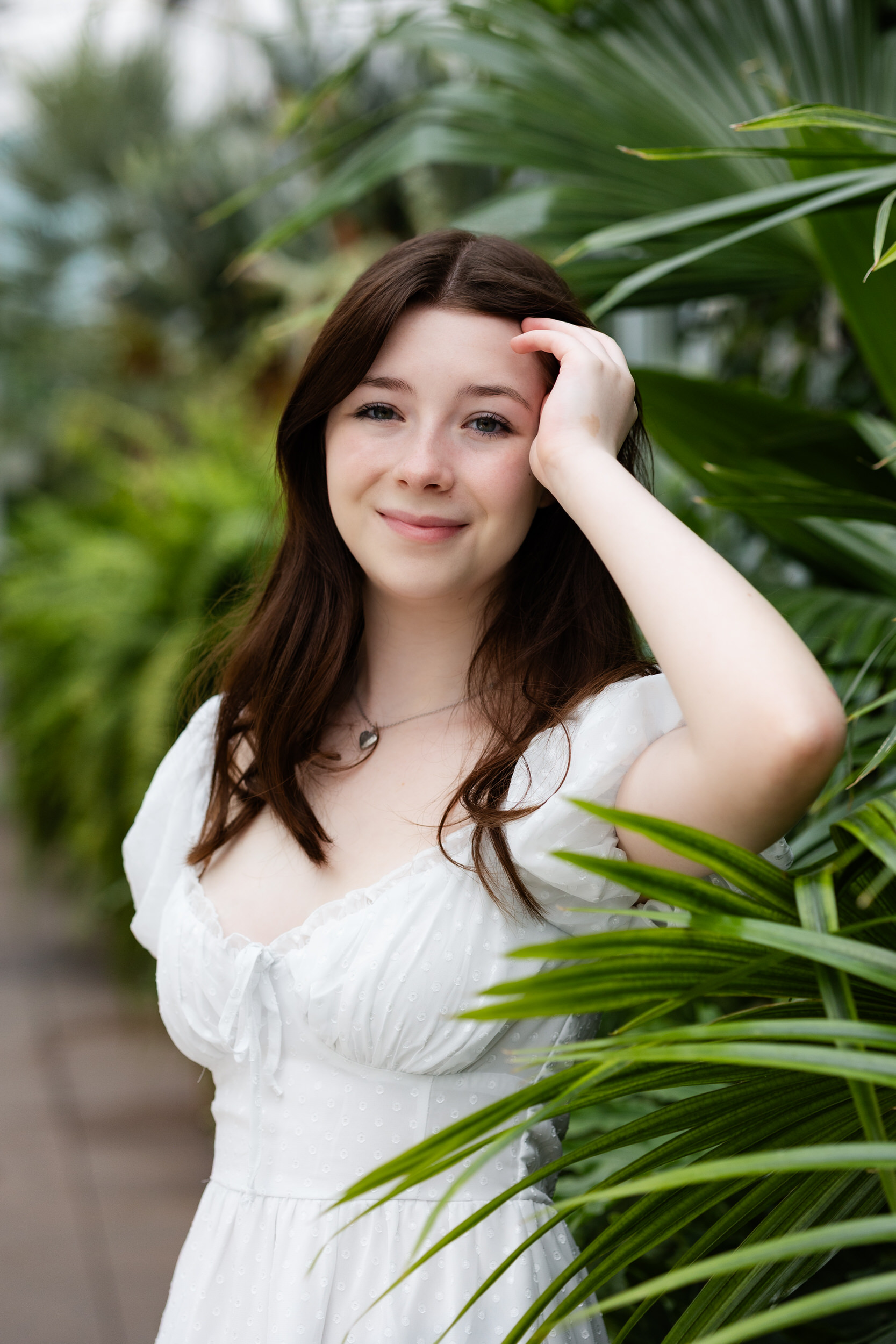 A young woman stands in a greenhouse and smiles at the camera while running one hand through her hair.