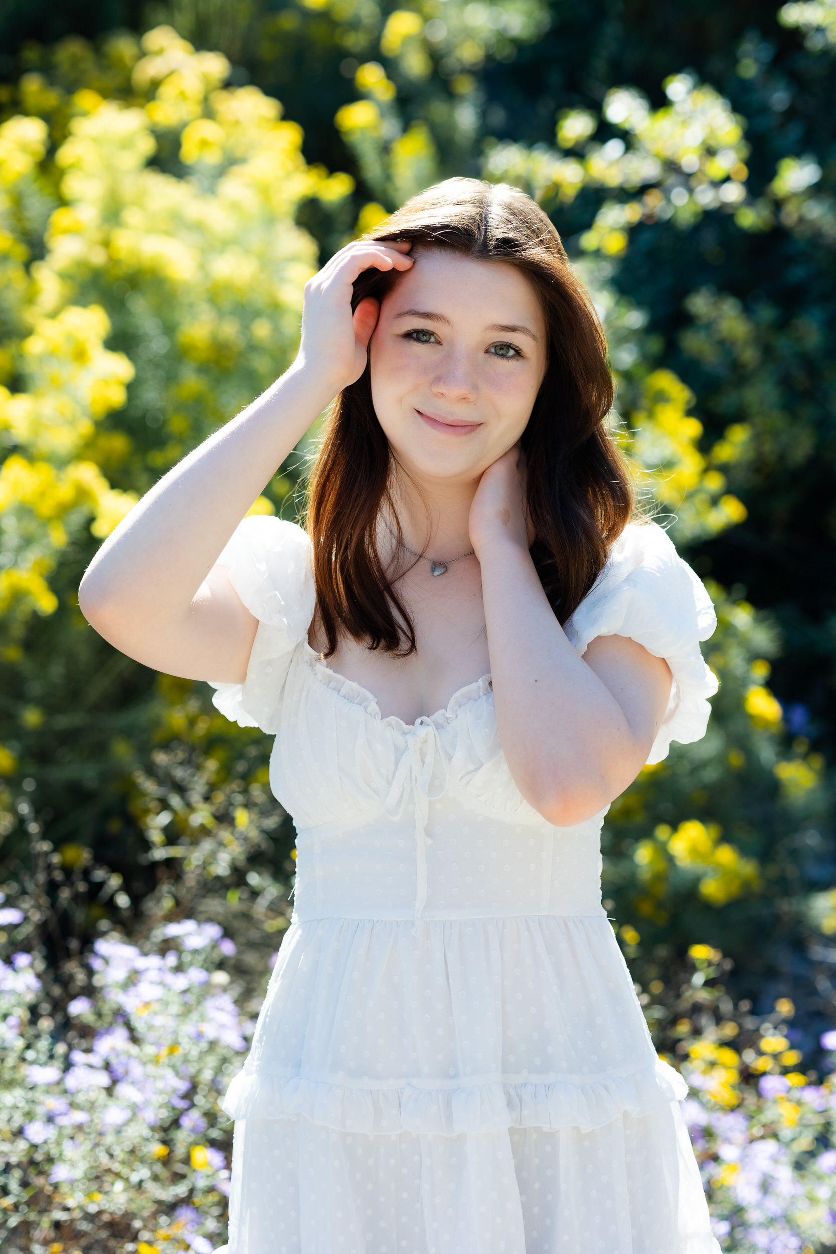 A young woman rests one hand on her neck and runs the other through her hair as she smiles at the camera.