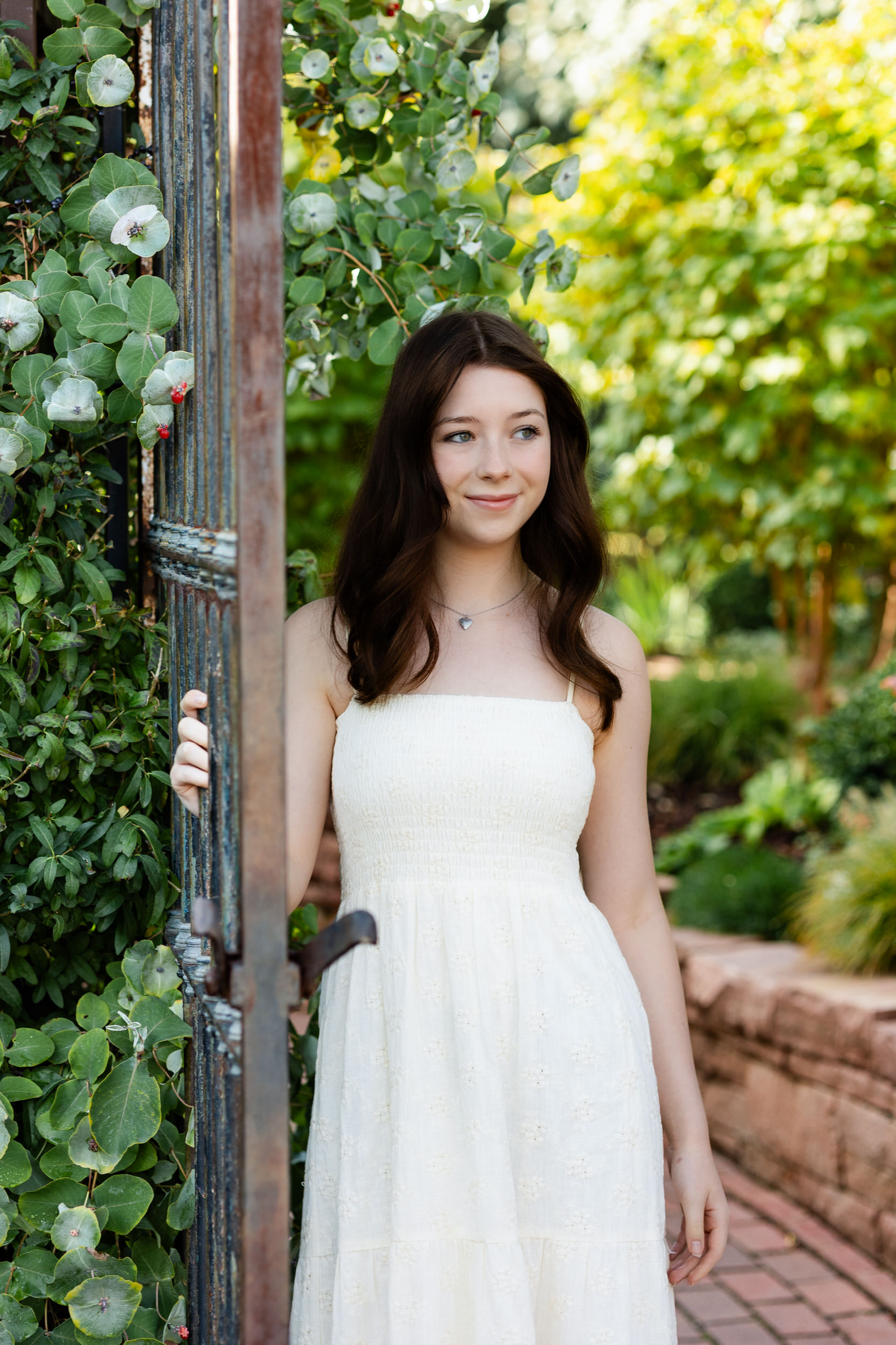 A young woman holds onto a garden gate with one hand and looks off-camera.