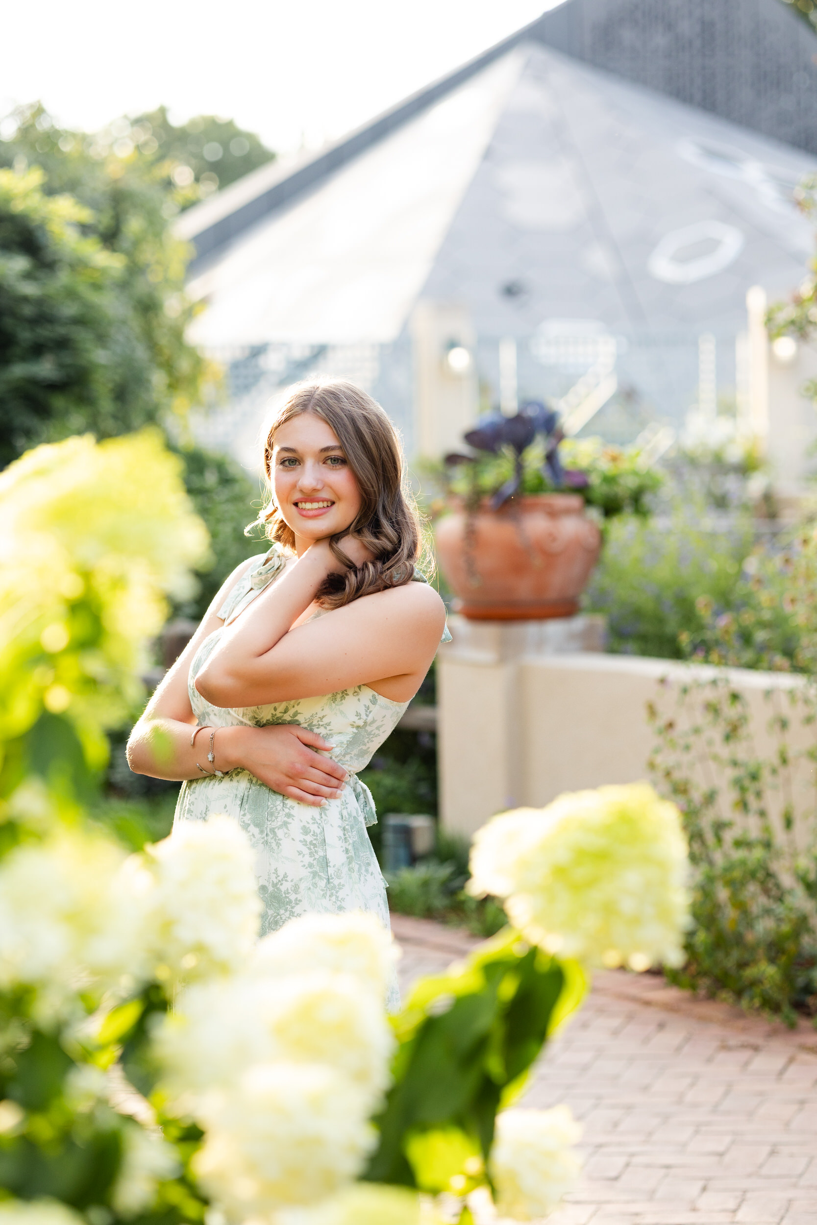 A young woman stands behind a bush with yellow flowers and smiles at the camera.