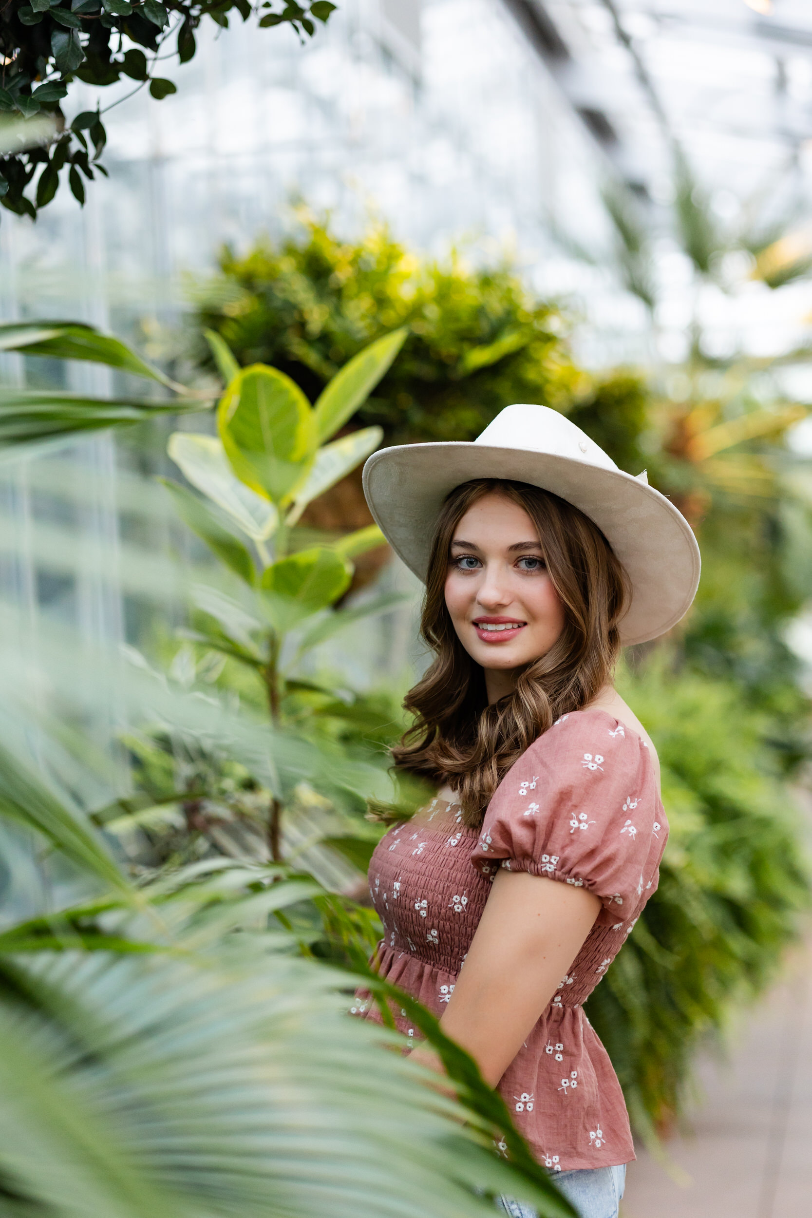 A young woman in a wide-brimmed hat stands in a greenhouse and smiles at the camera.