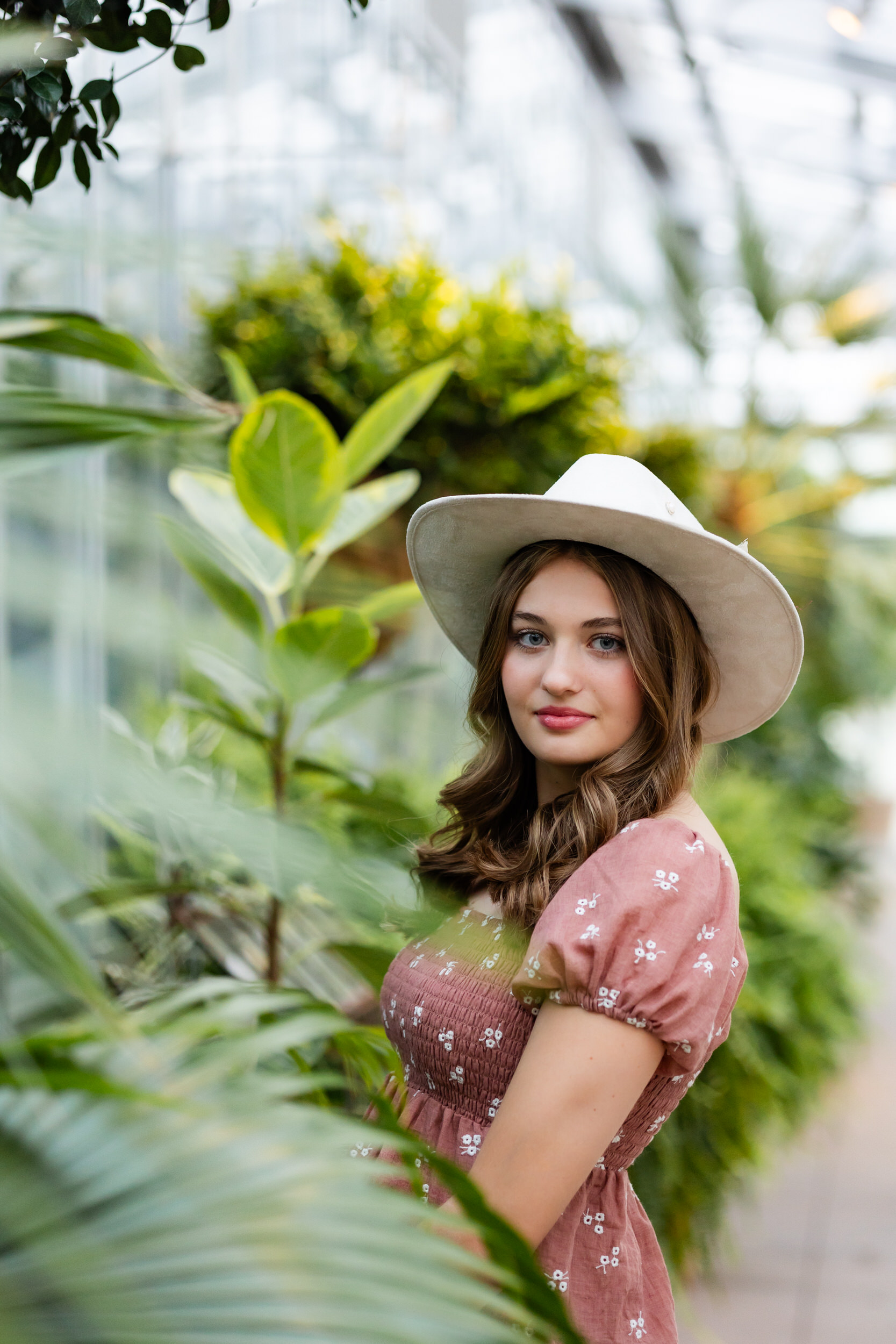 A young woman in a wide-brimmed hat stands in a greenhouse and smiles at the camera.