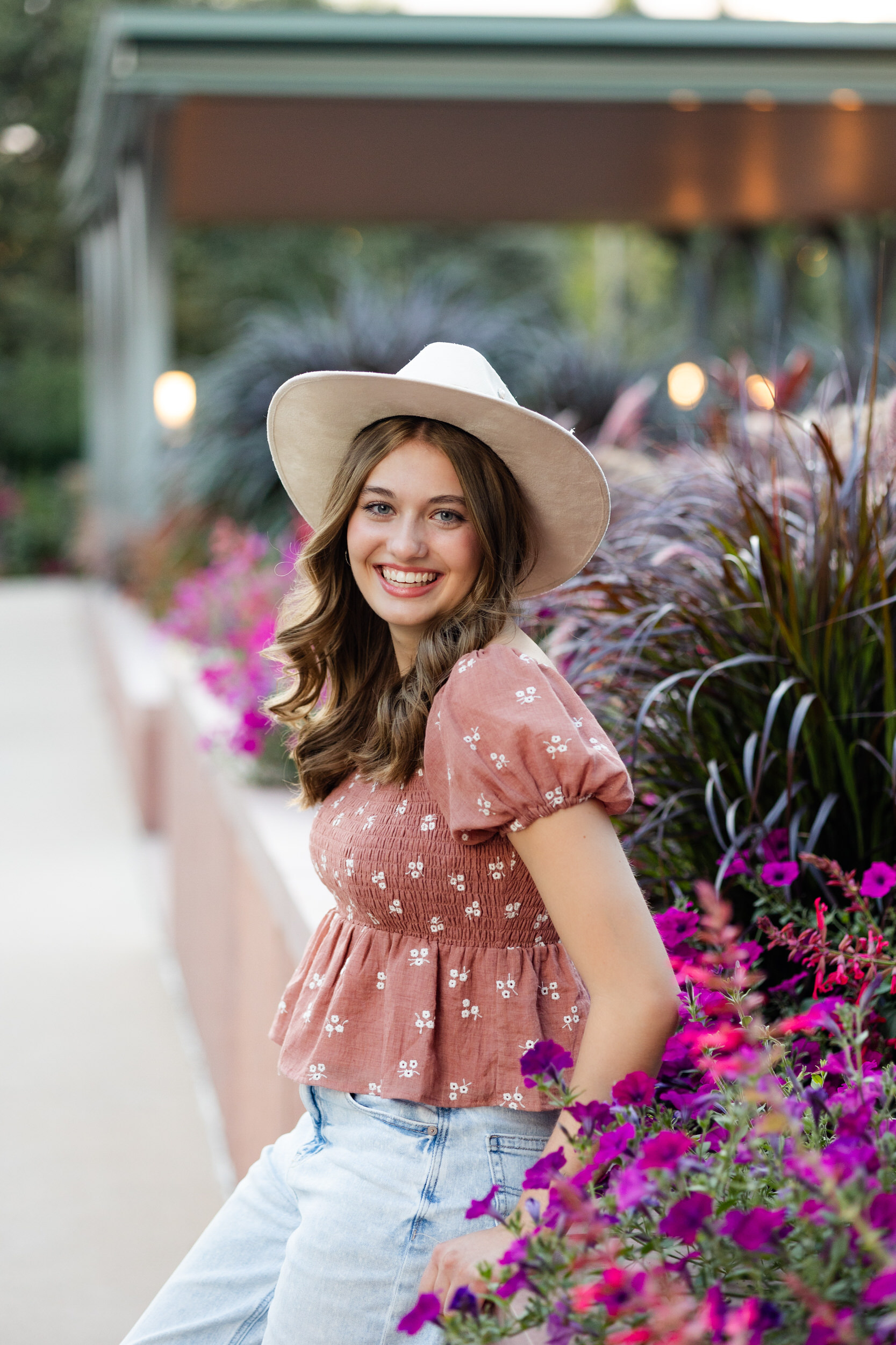 A young woman in a wide-brimmed hat sits on a garden ledge and smiles.