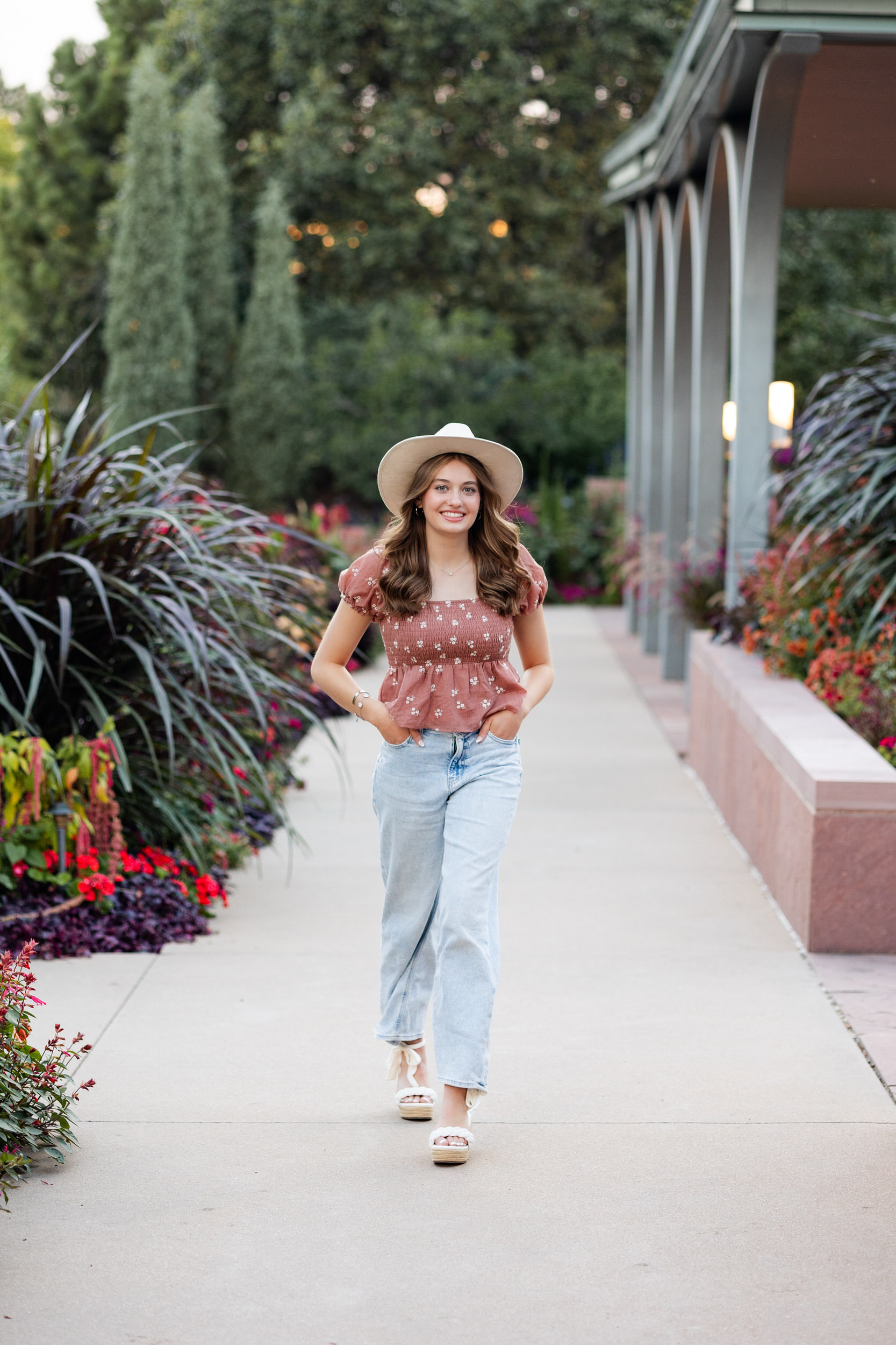A young woman in a wide-brimmed hat walks toward the camera and smiles.