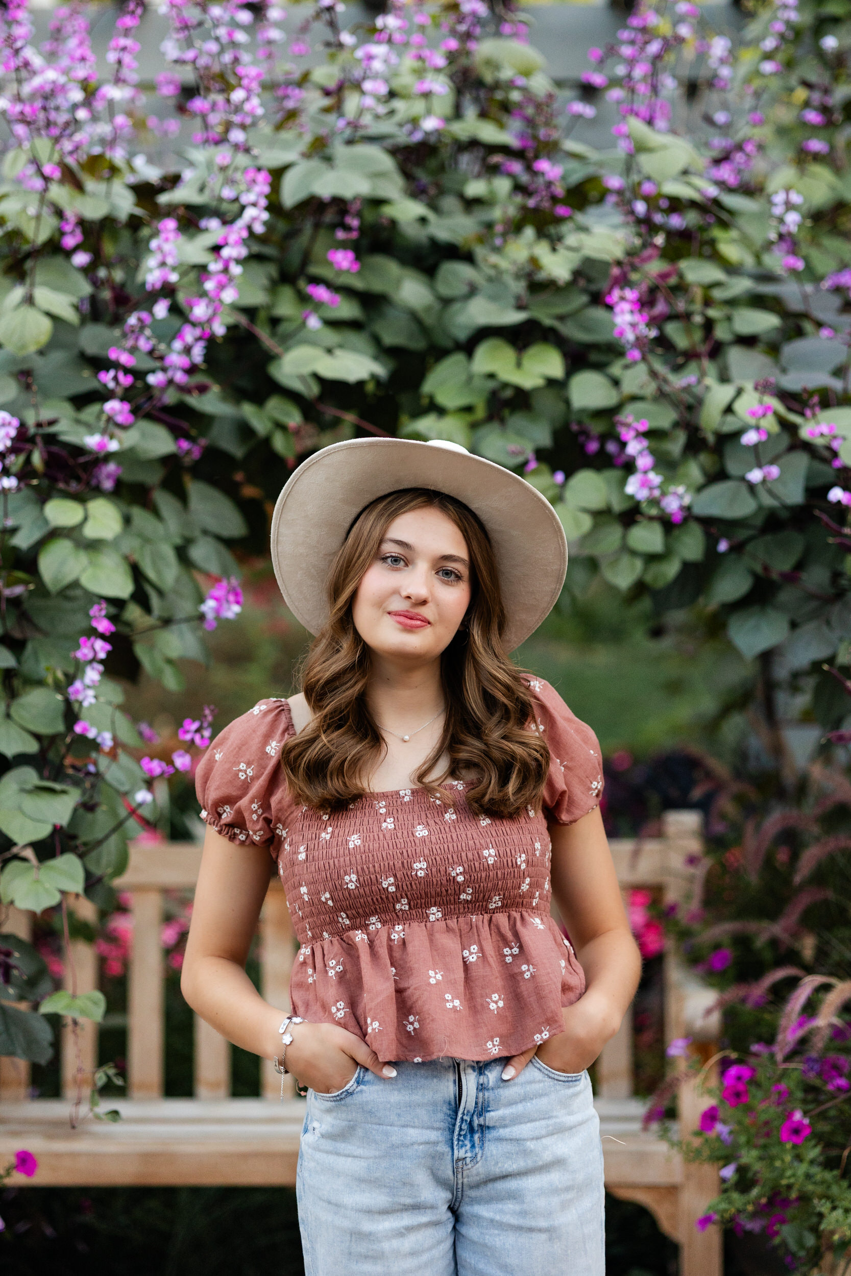 A young woman in a wide-brimmed hat is surrounded by hanging purples flowers and smiles at the camera.