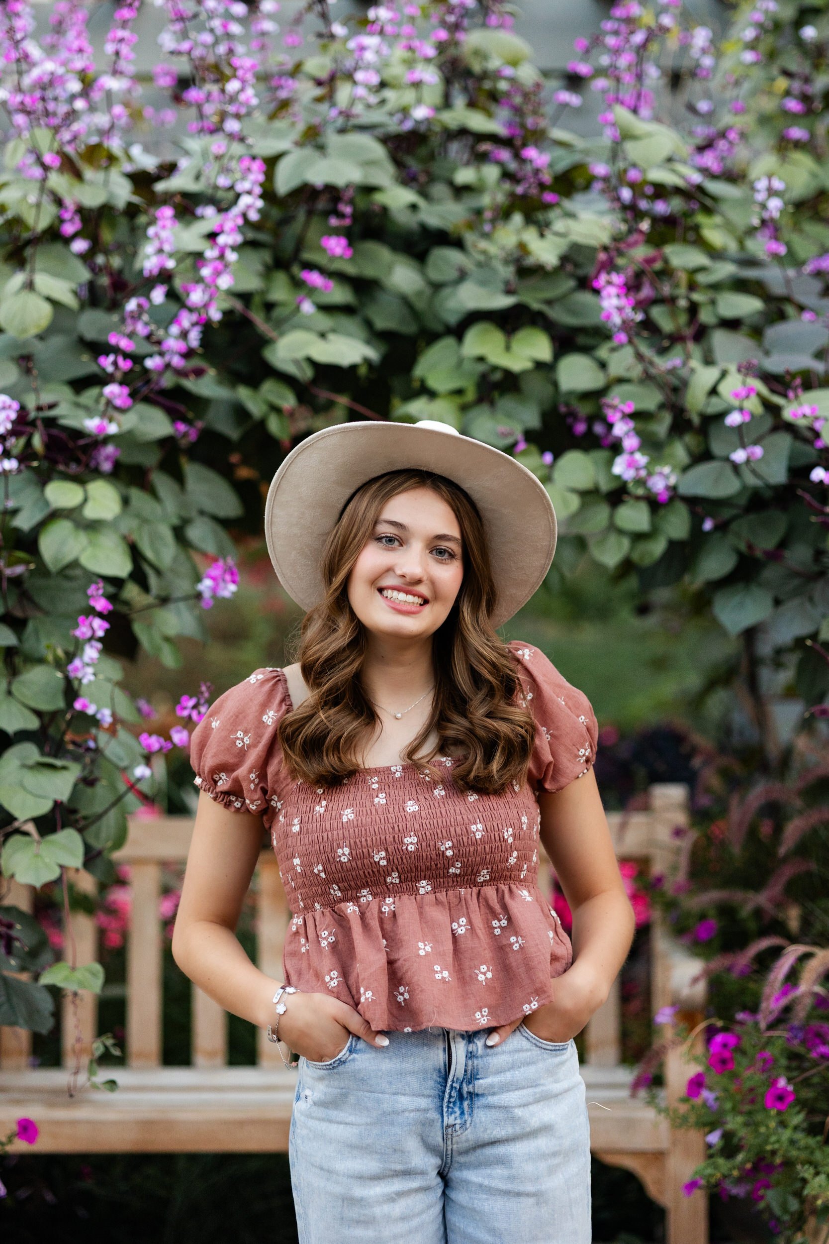 A young woman in a wide-brimmed hat is surrounded by hanging purples flowers and smiles at the camera.