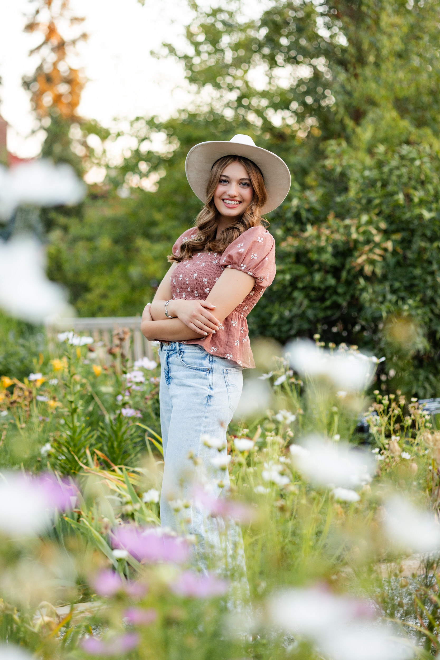 A young woman in a wide-brimmed hat stands in a garden and smiles at the camera.