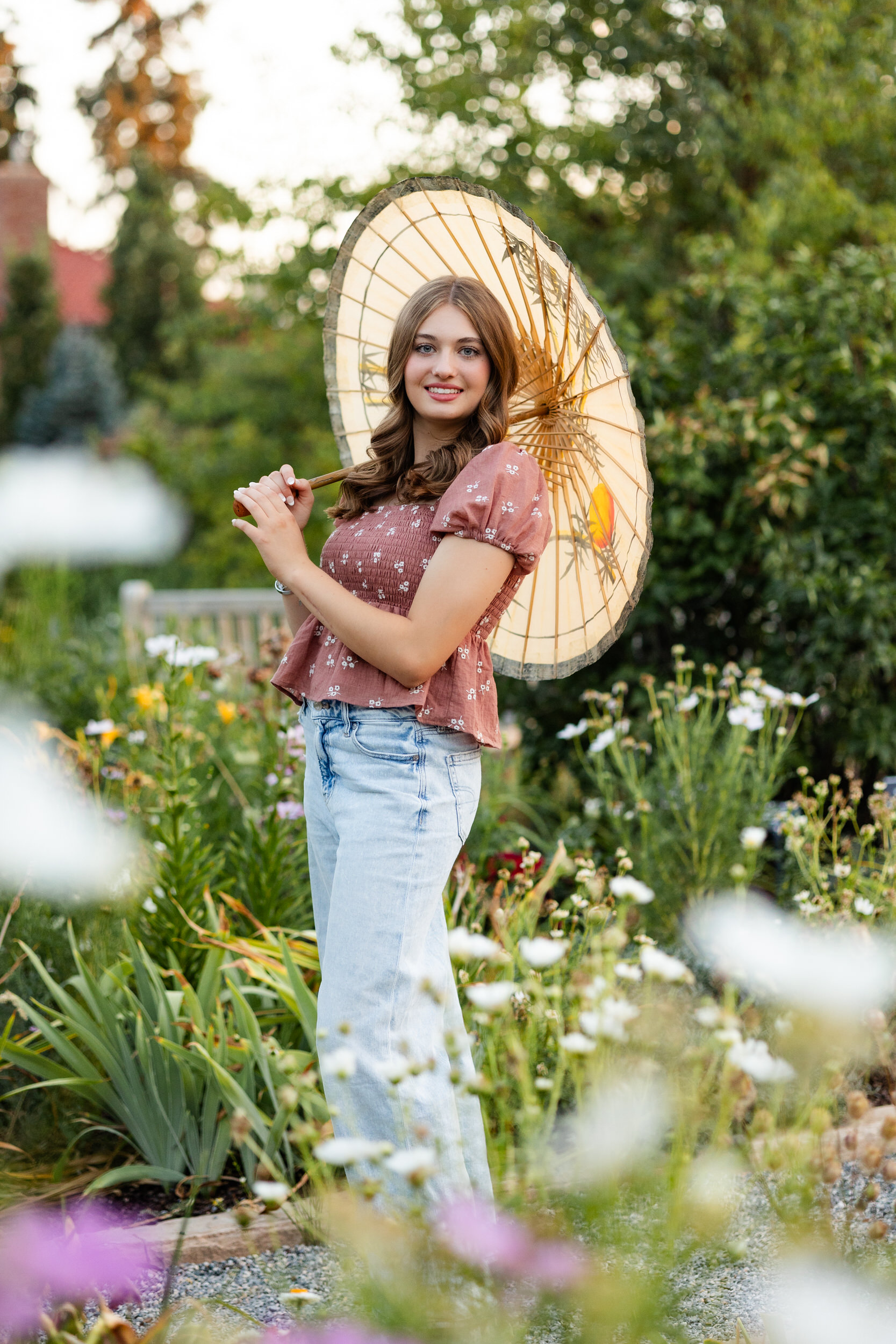A young woman stands in a garden with a parasol on one shoulder and smiles at the camera.