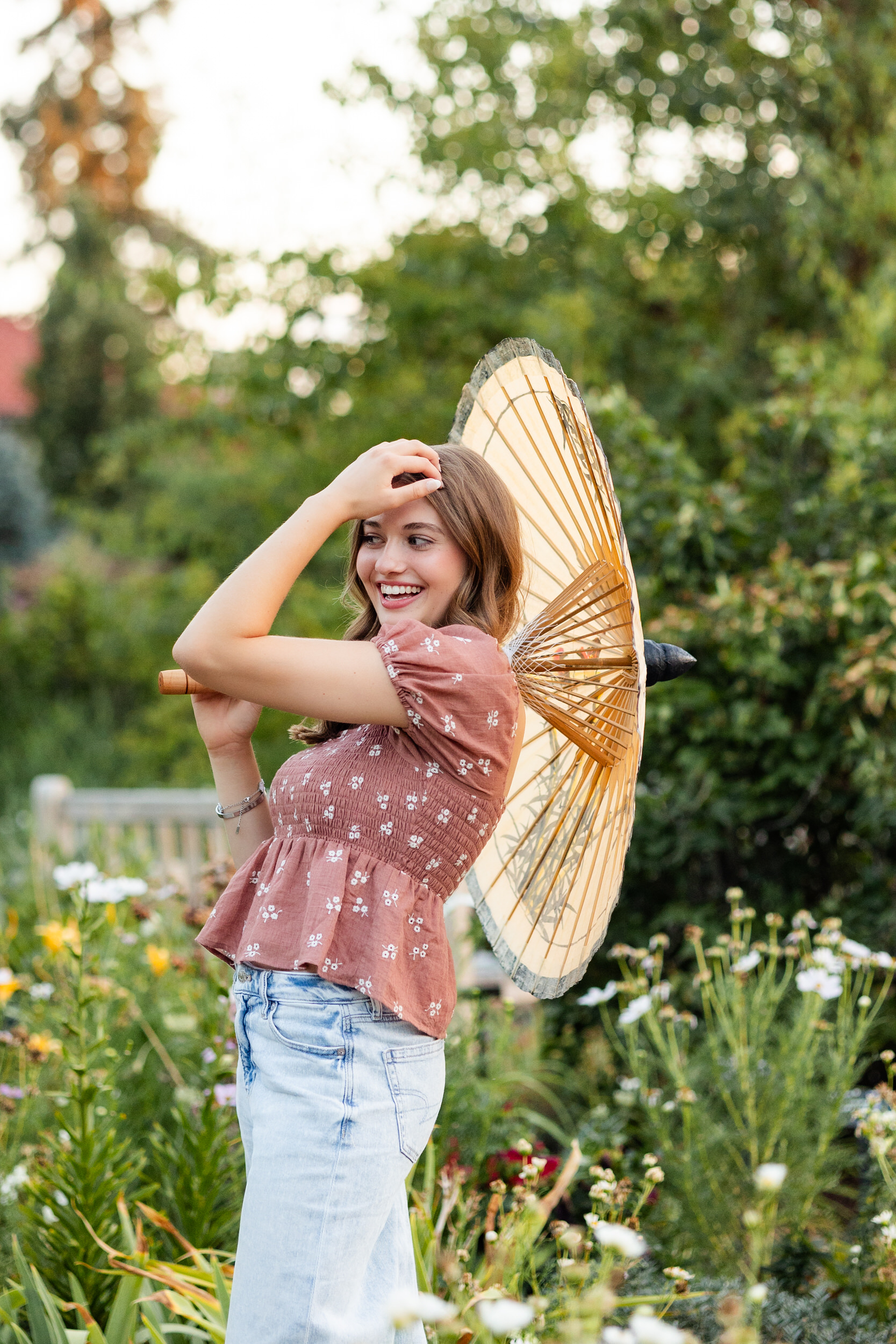 A young woman stands in a garden with a parasol on one shoulder and smiles off camera.