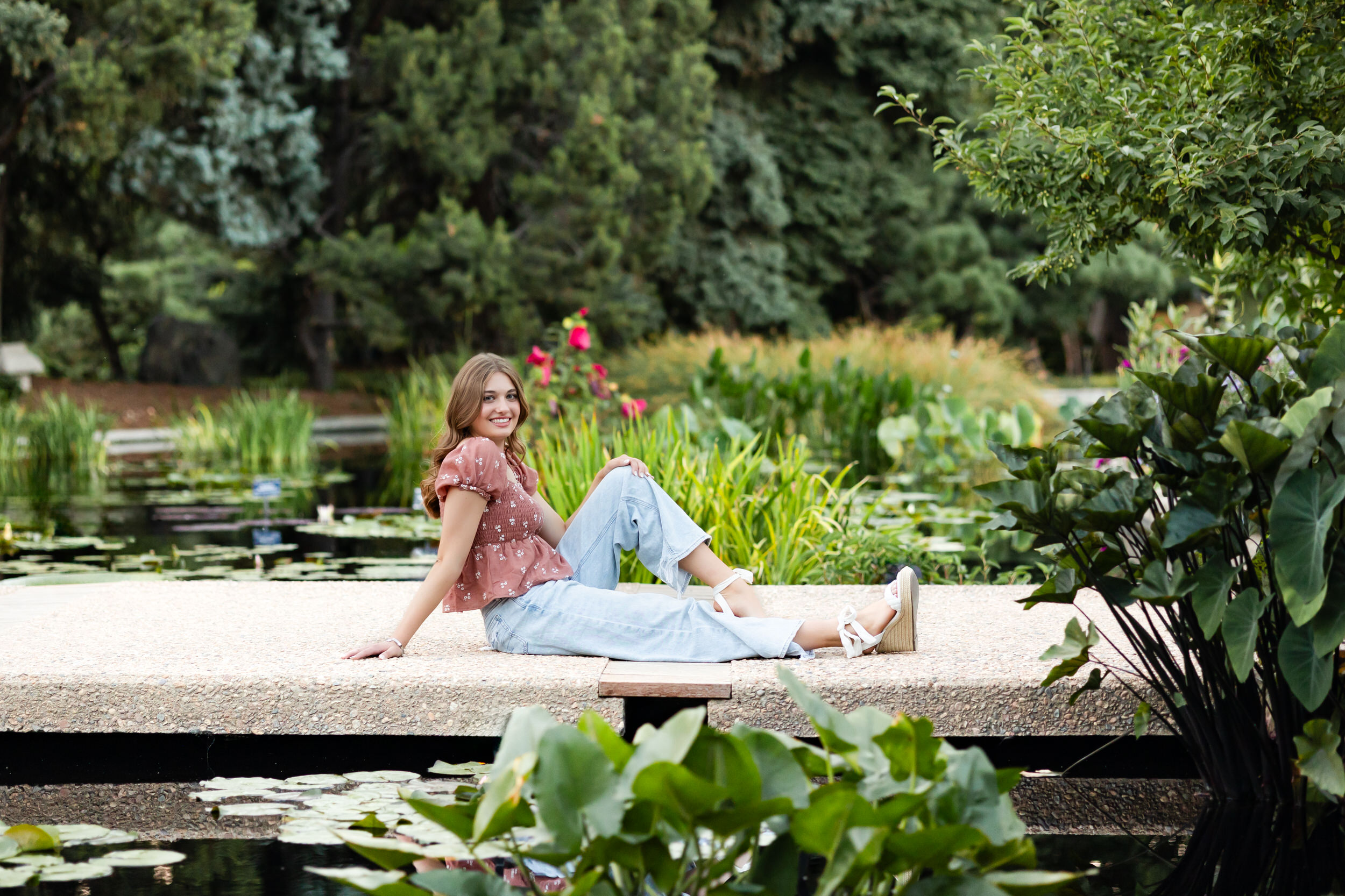 A young woman sits on a bridge over a pond and smiles at the camera.