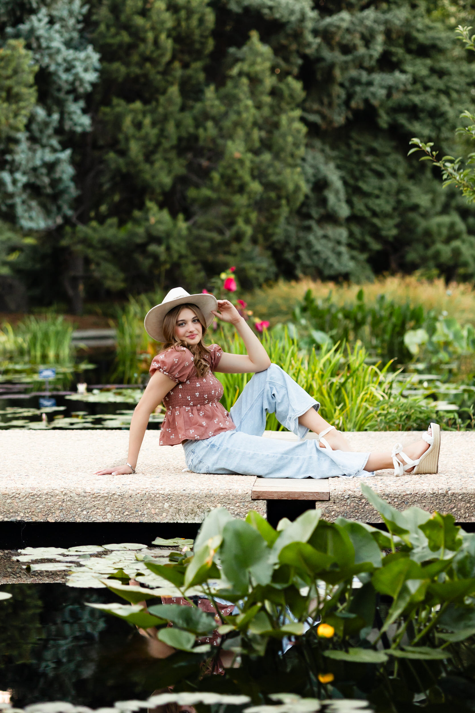 A young woman sits on a bridge over a pond and smiles at the camera.