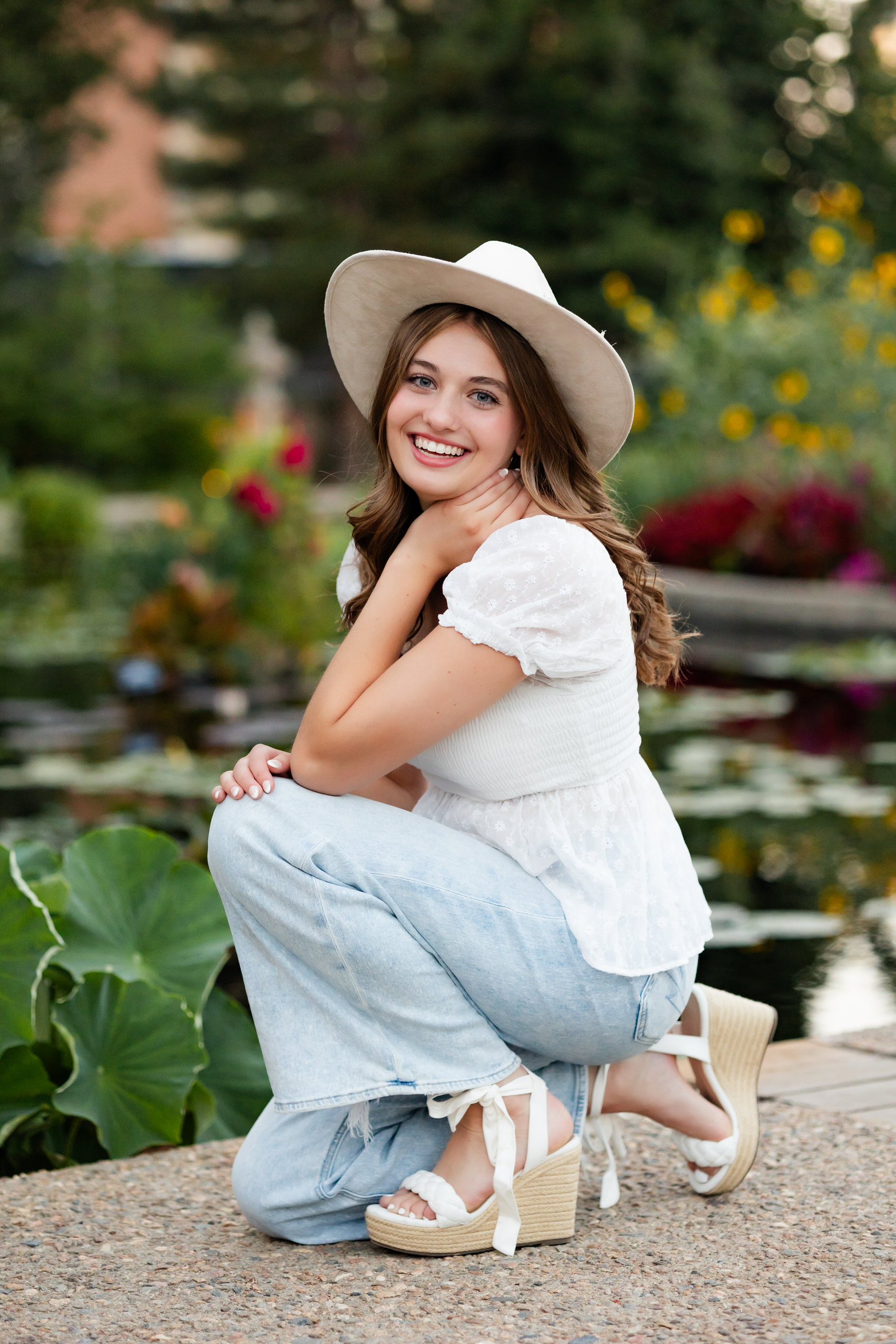 A young woman in a wide-brimmed hat squats down near a pond and smiles at the camera.