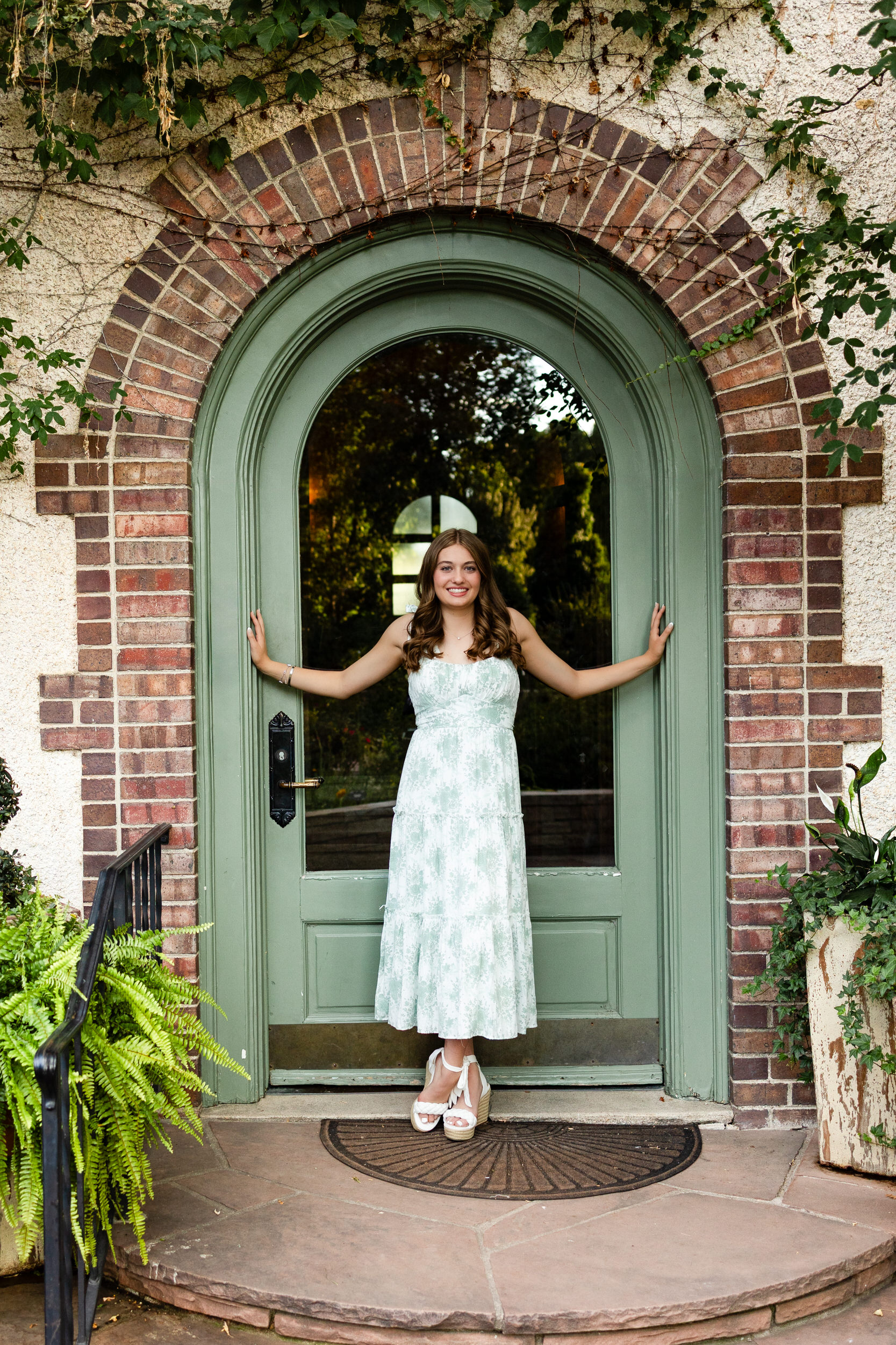 A young woman in a white and green dress stands in a garden doorway and smiles at the camera.