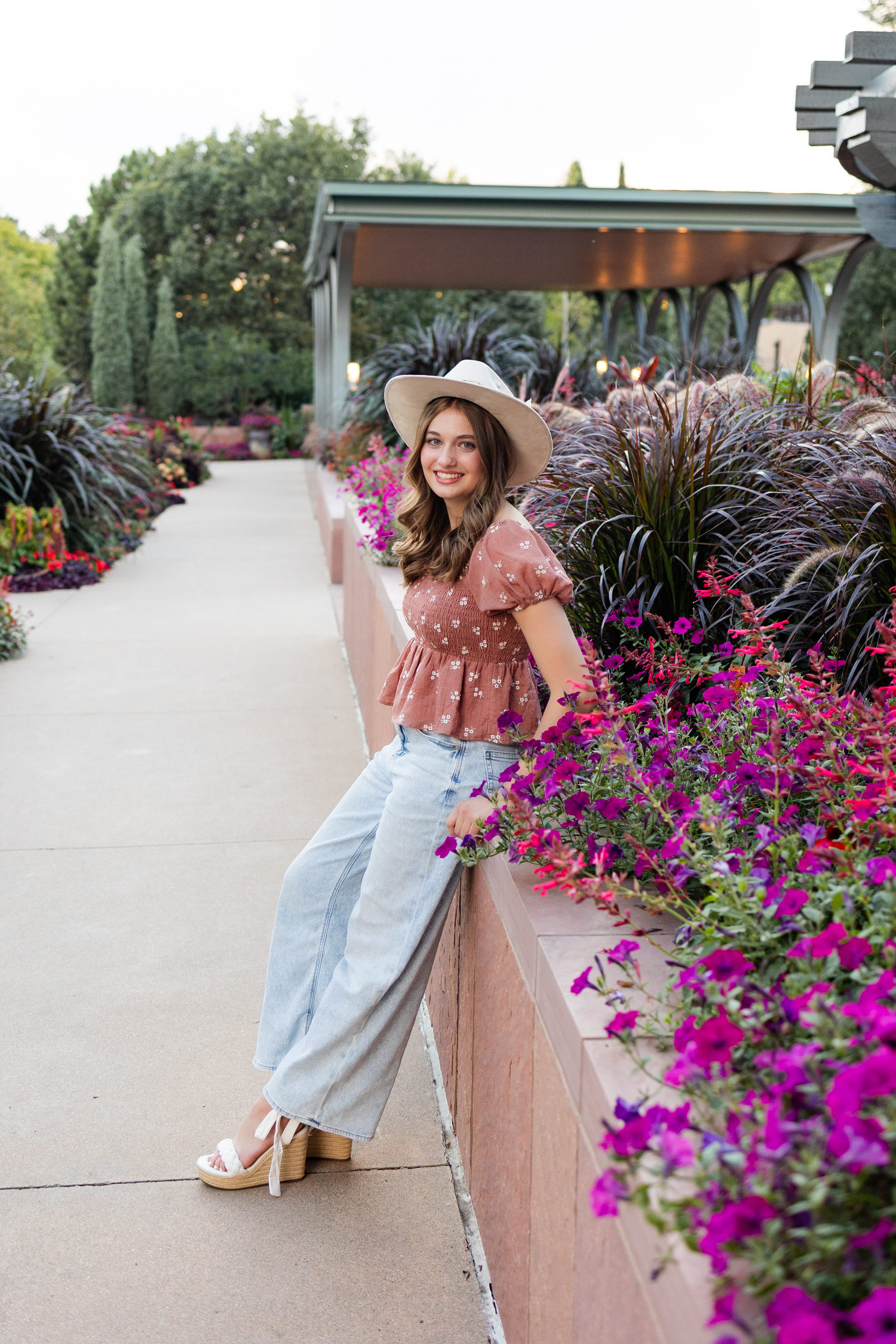 A young woman sits on a ledge in a garden and smiles at the camera.