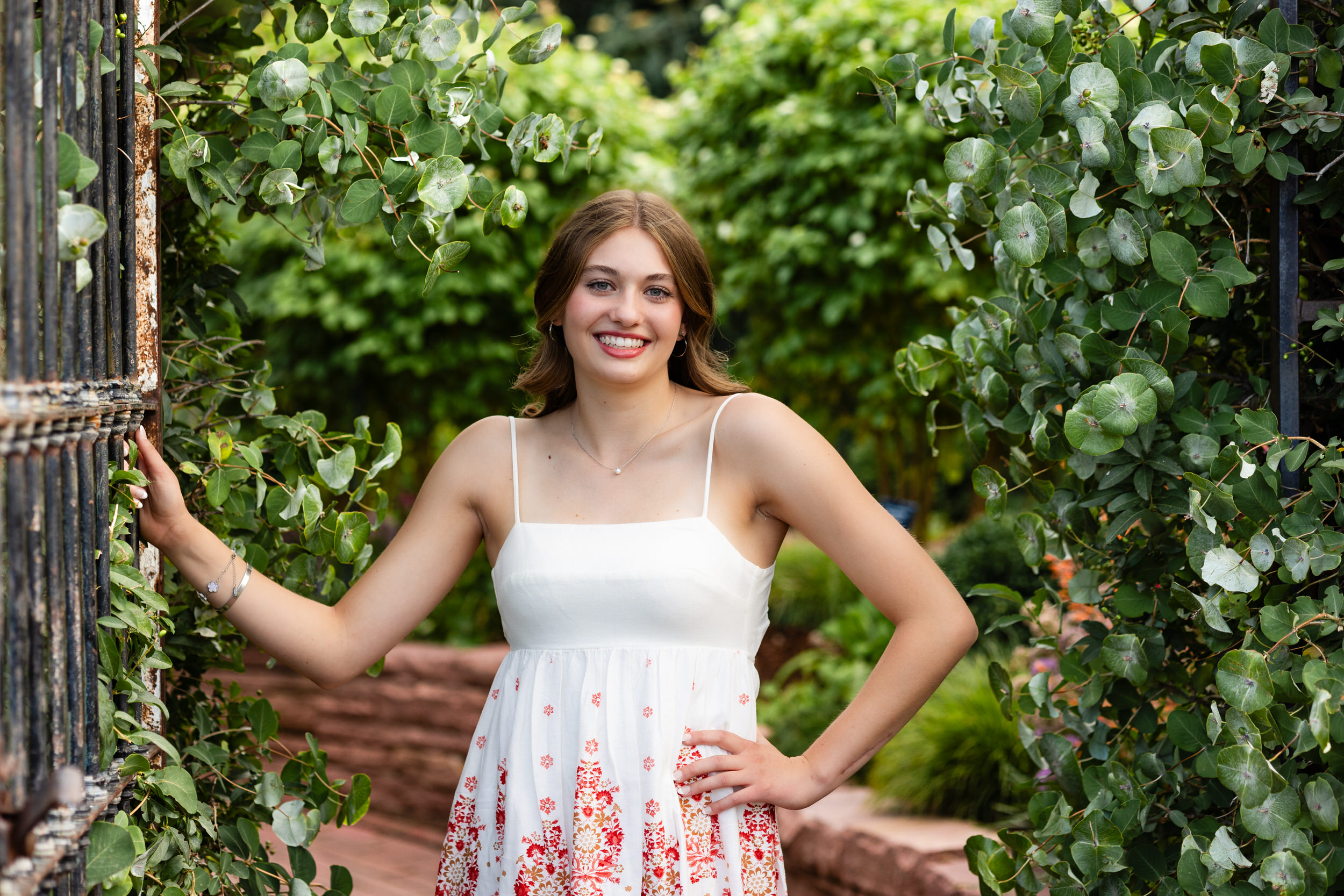A young woman stands with one hand on a garden gate and one on her waist and smiles at the camera.