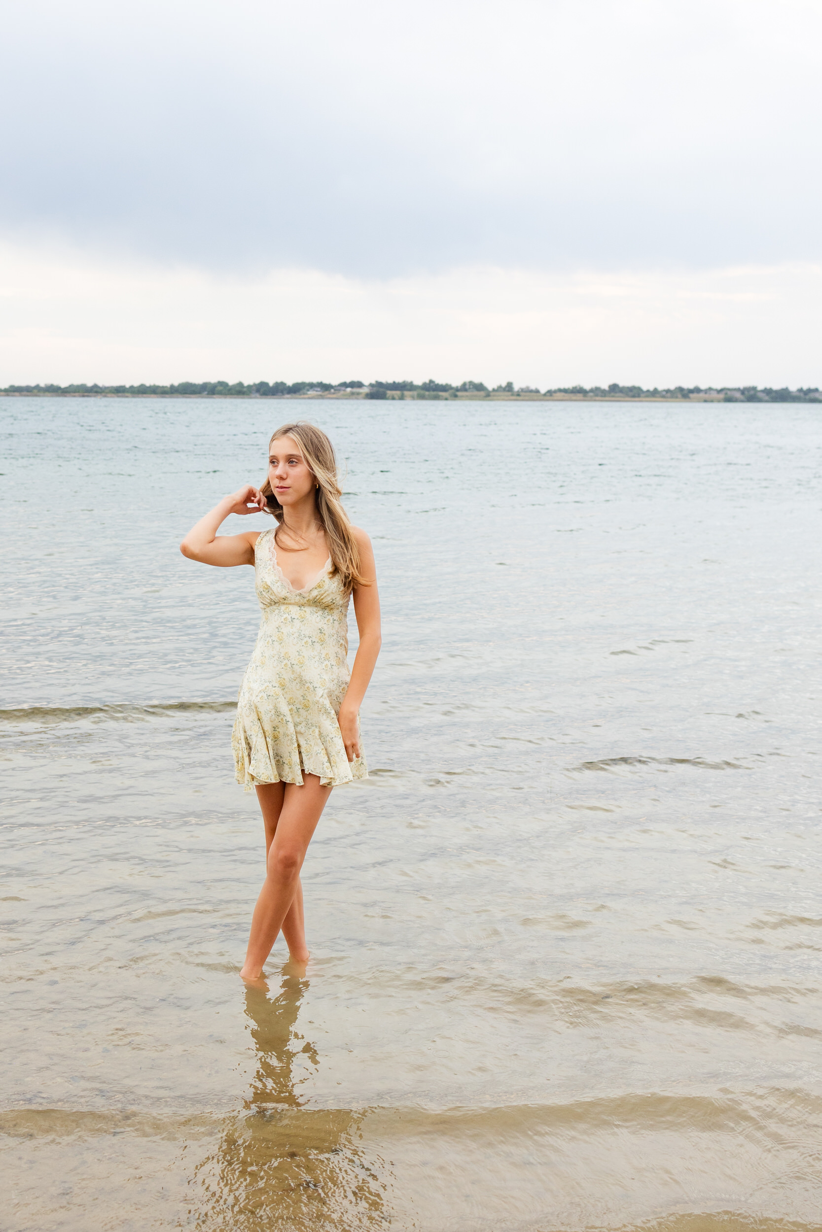 A young woman stands ankle-deep in a lake and runs her hand through her hair as she looks off-camera.