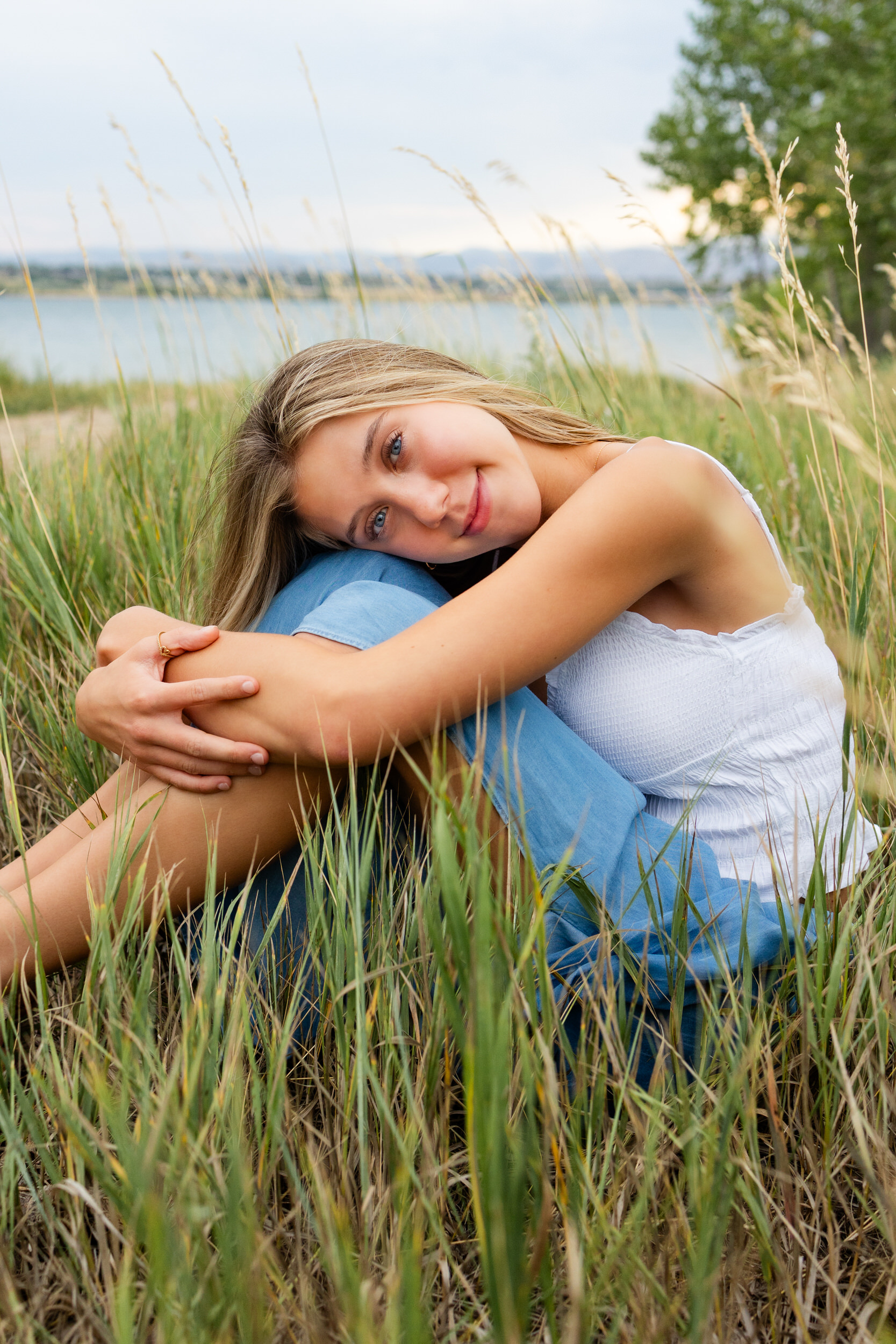 A young woman sits in tall grass hugging her knees.
