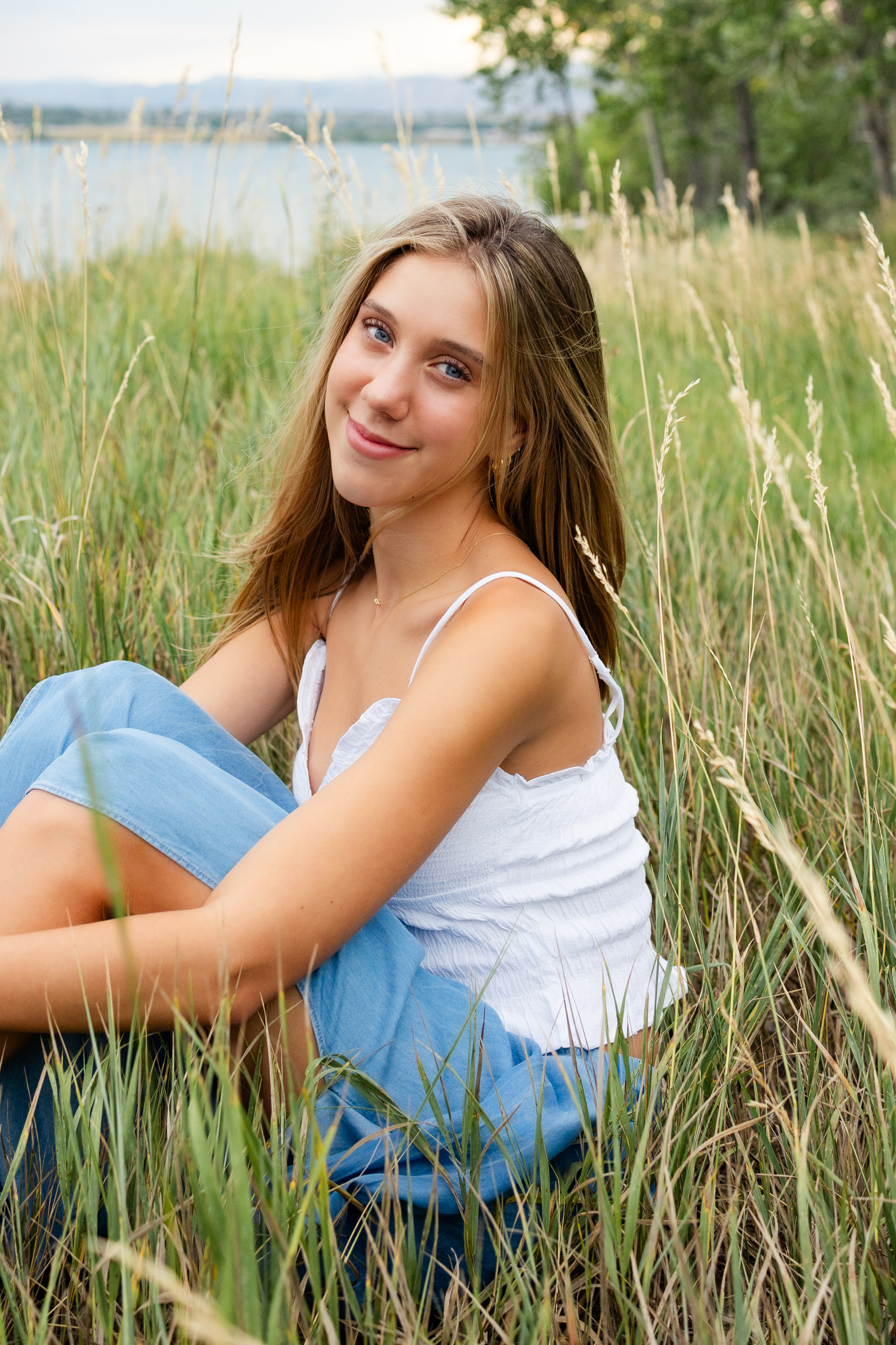 A young woman sits in tall grass hugging her knees and smiles at the camera.