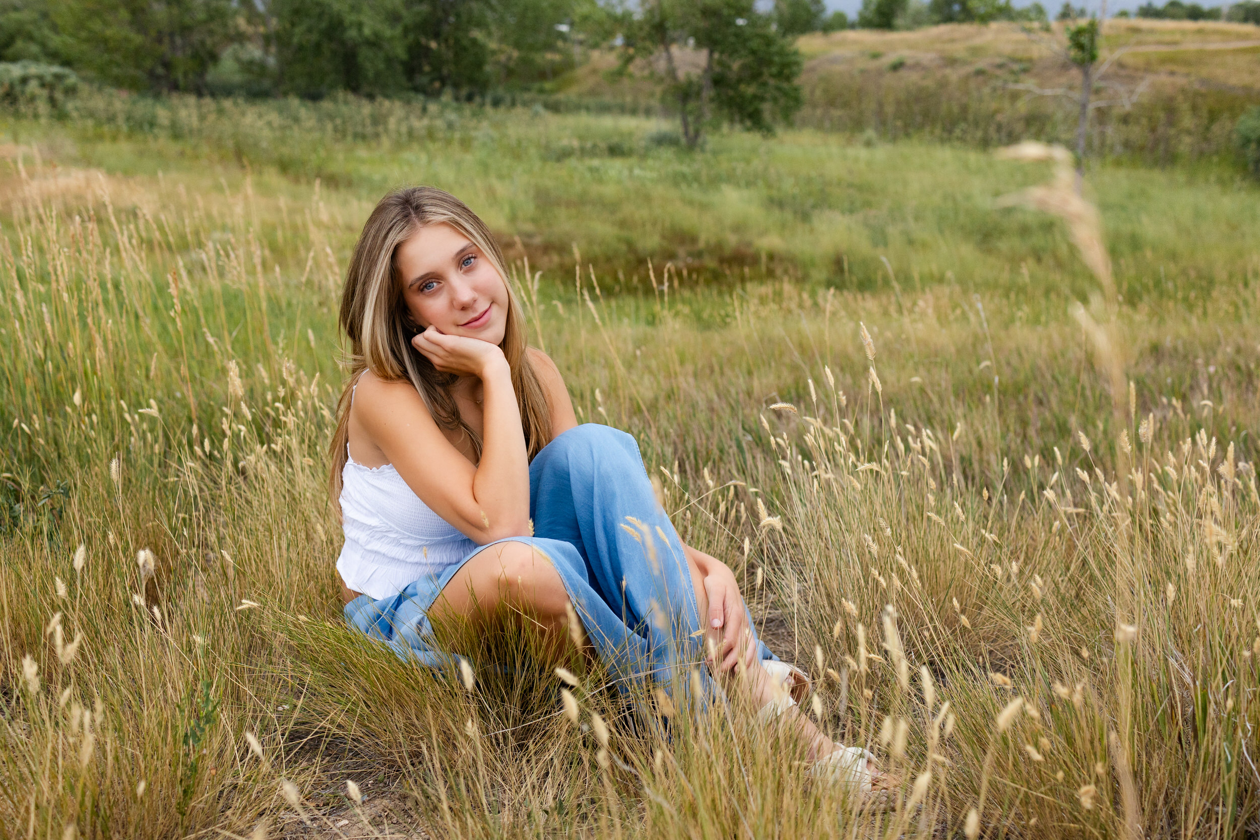 A young woman sits in a field of tall grass hugging one knee and smiles at the camera.
