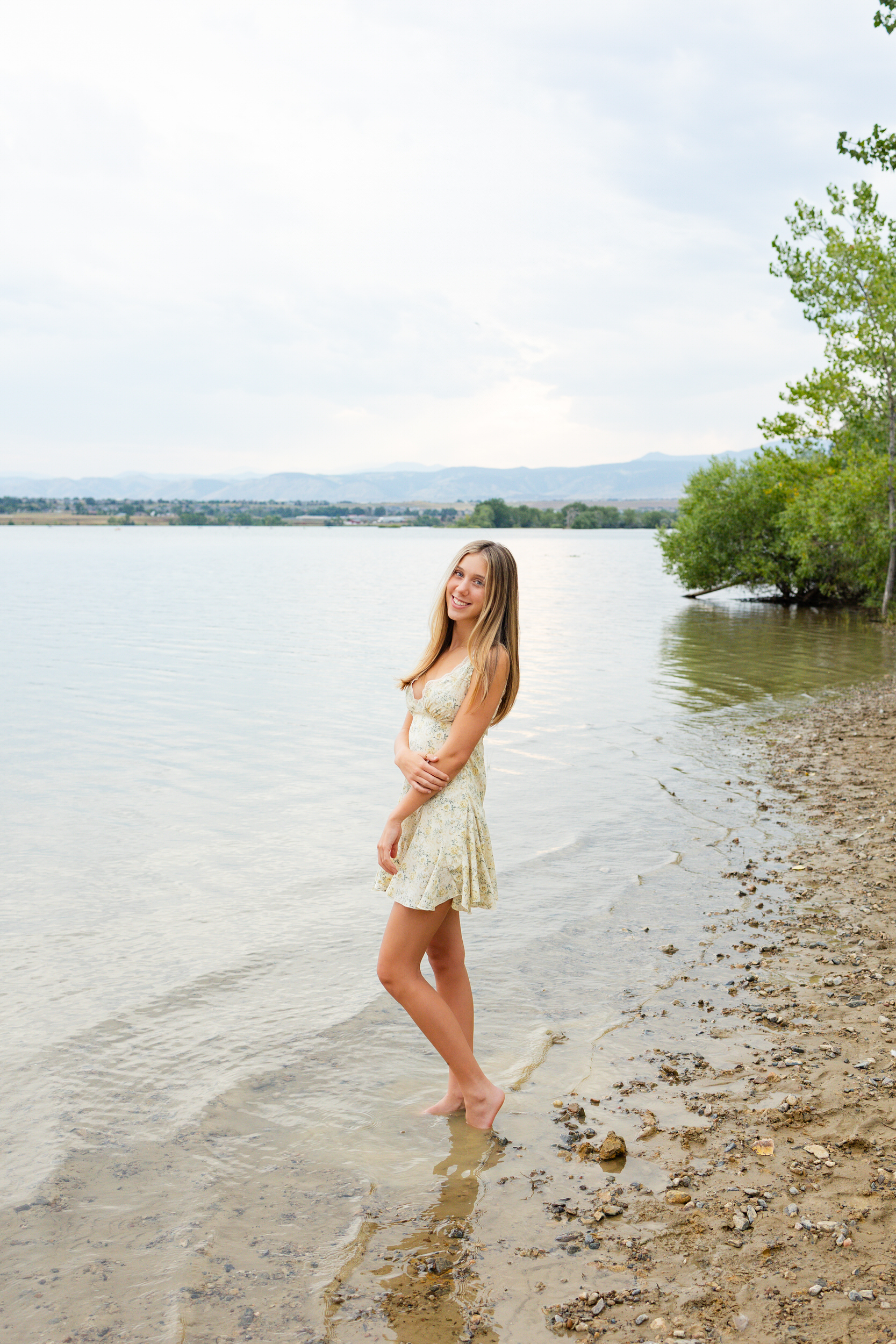 A young woman stands barefoot in a lake and smiles at the camera.