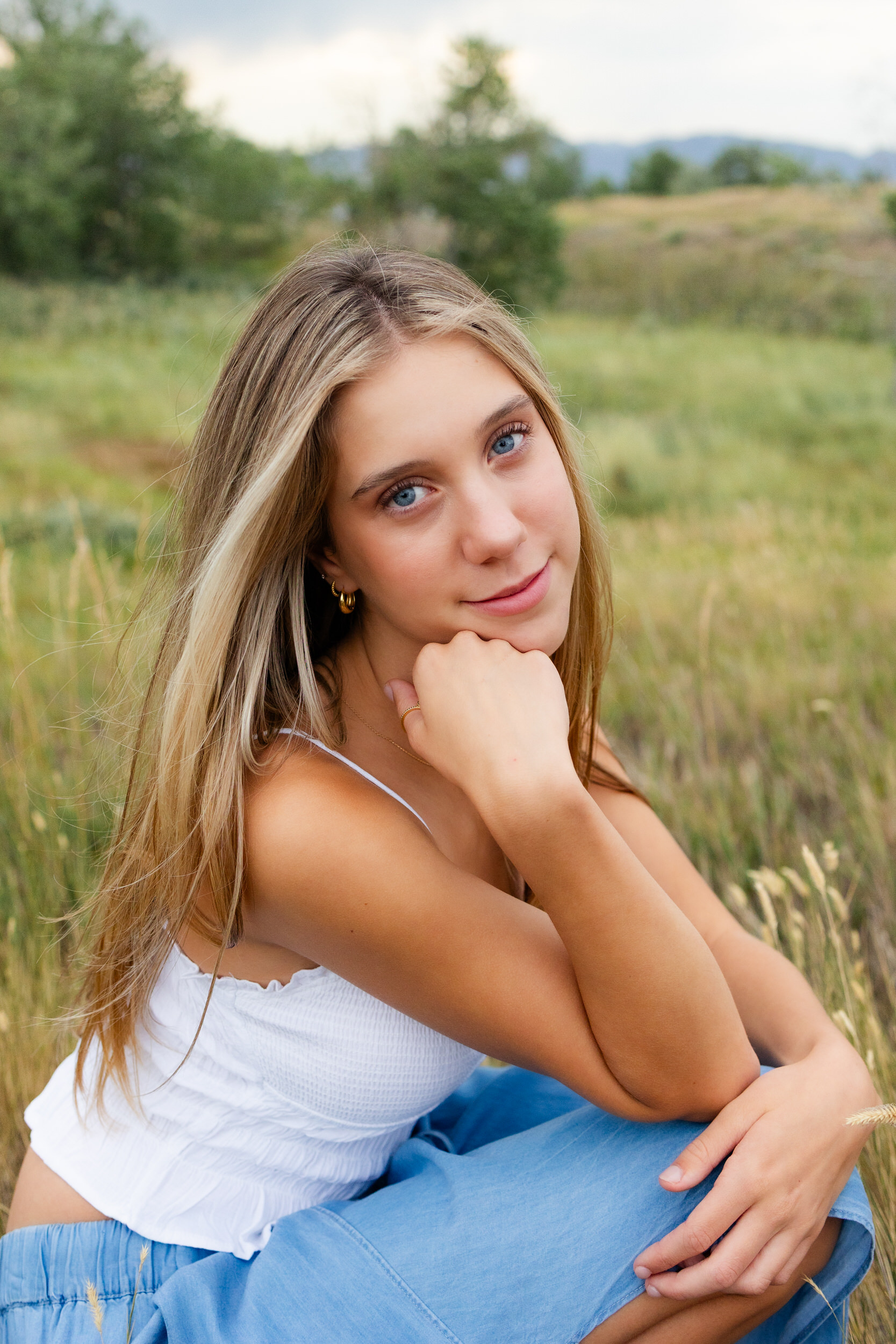 A young woman sits in a field and rests her chin in her hand and smiles at the camera.