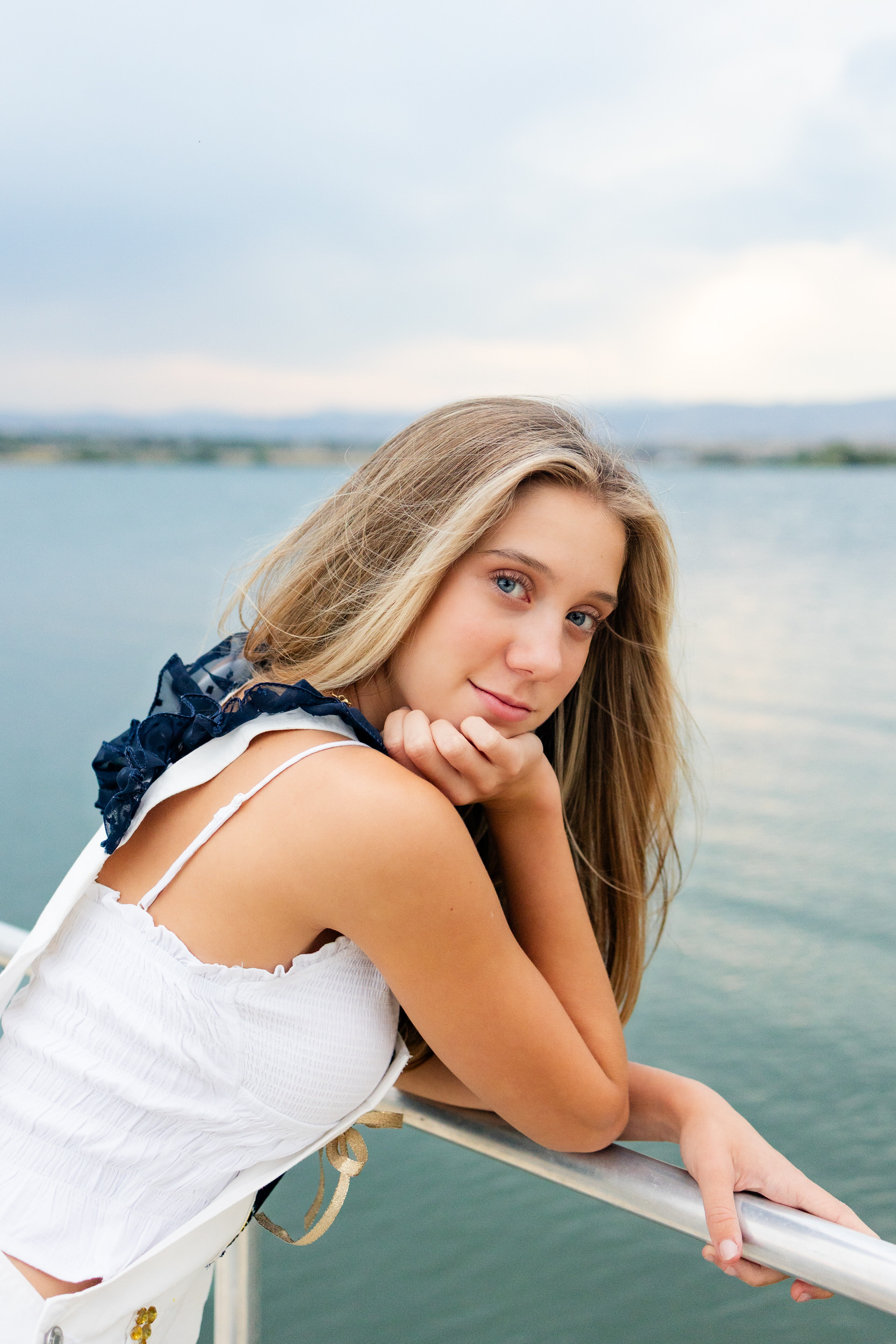 A young woman in senior overalls leans on a dock and looks over her shoulder to smile at the camera.