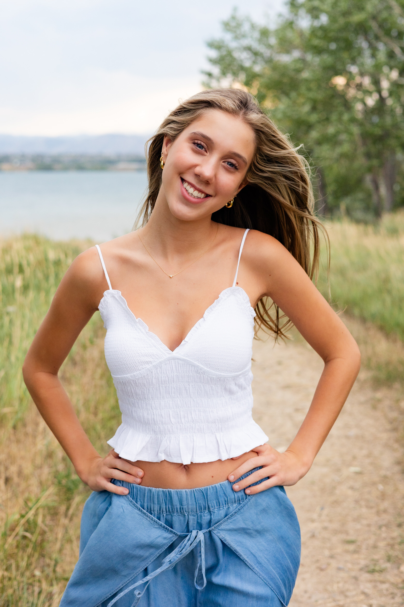 A young woman stands on a dirt path lined with tall grass with her hands resting on her hips and smiles at the camera.
