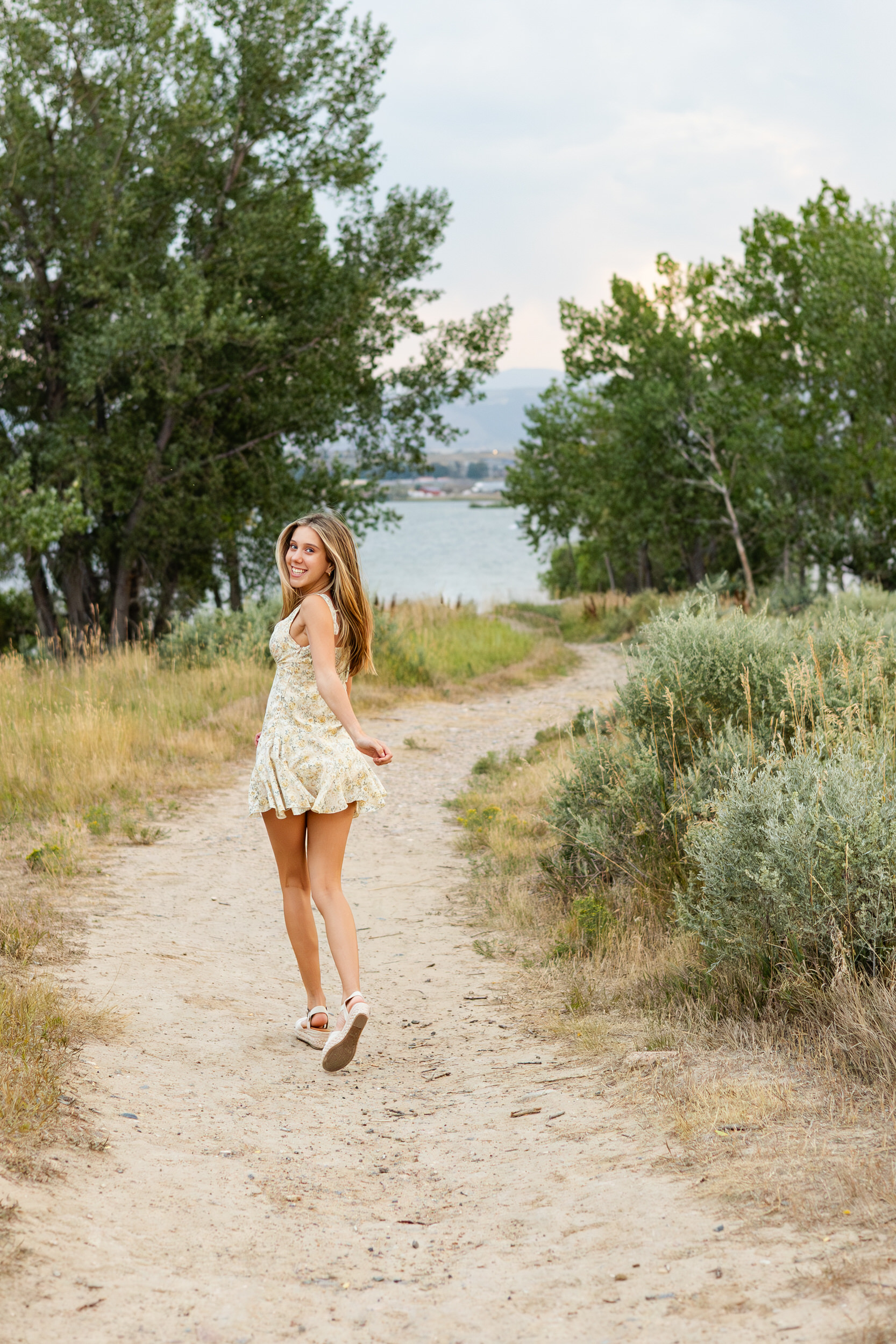 A young woman in a short floral dress runs down a dirt path lined by tall grass and looks over her shoulder smiling at the camera.