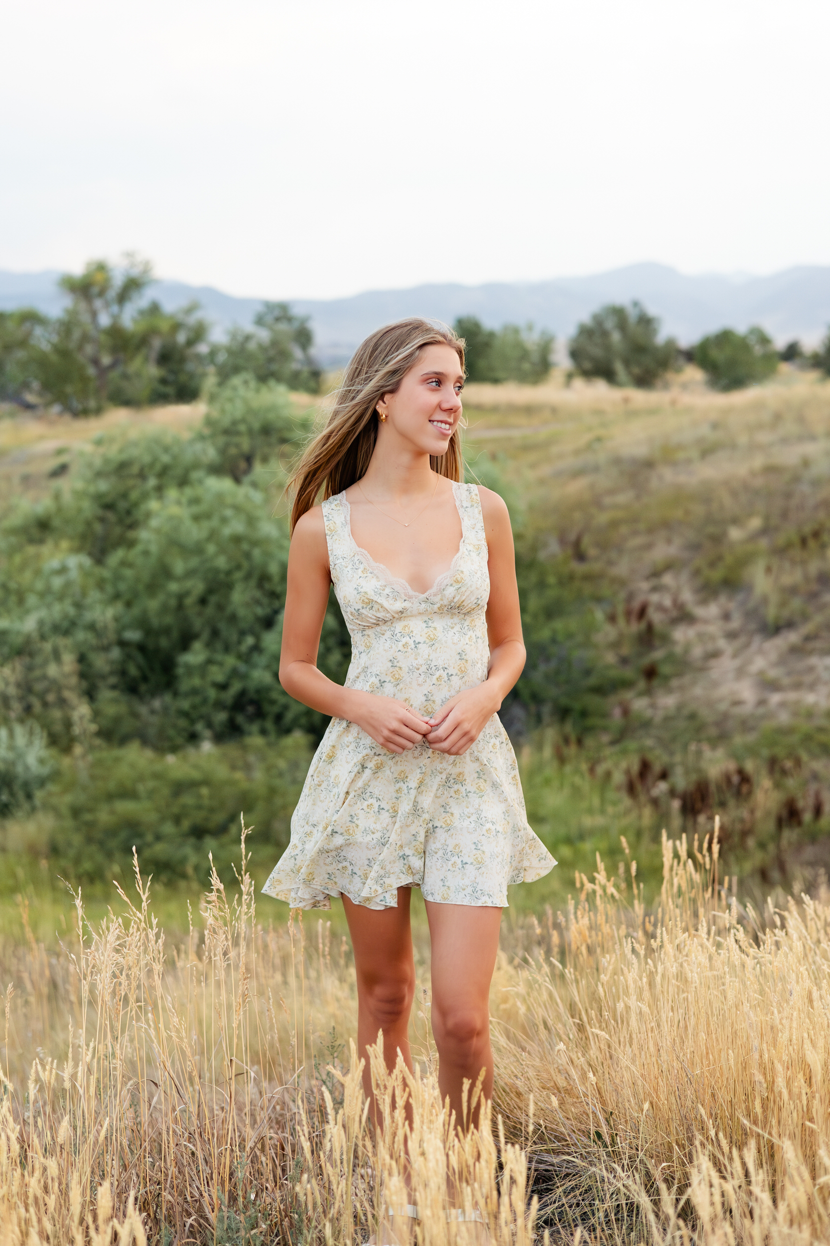 A young woman in a short floral dress stands in tall grass and smiles over her shoulder off-camera.