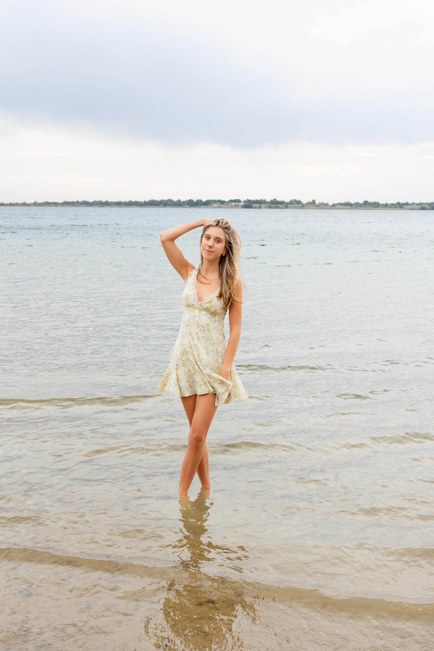 A young woman stands ankle-deep in a lake and smiles at the camera while she runs a hand through her hair.