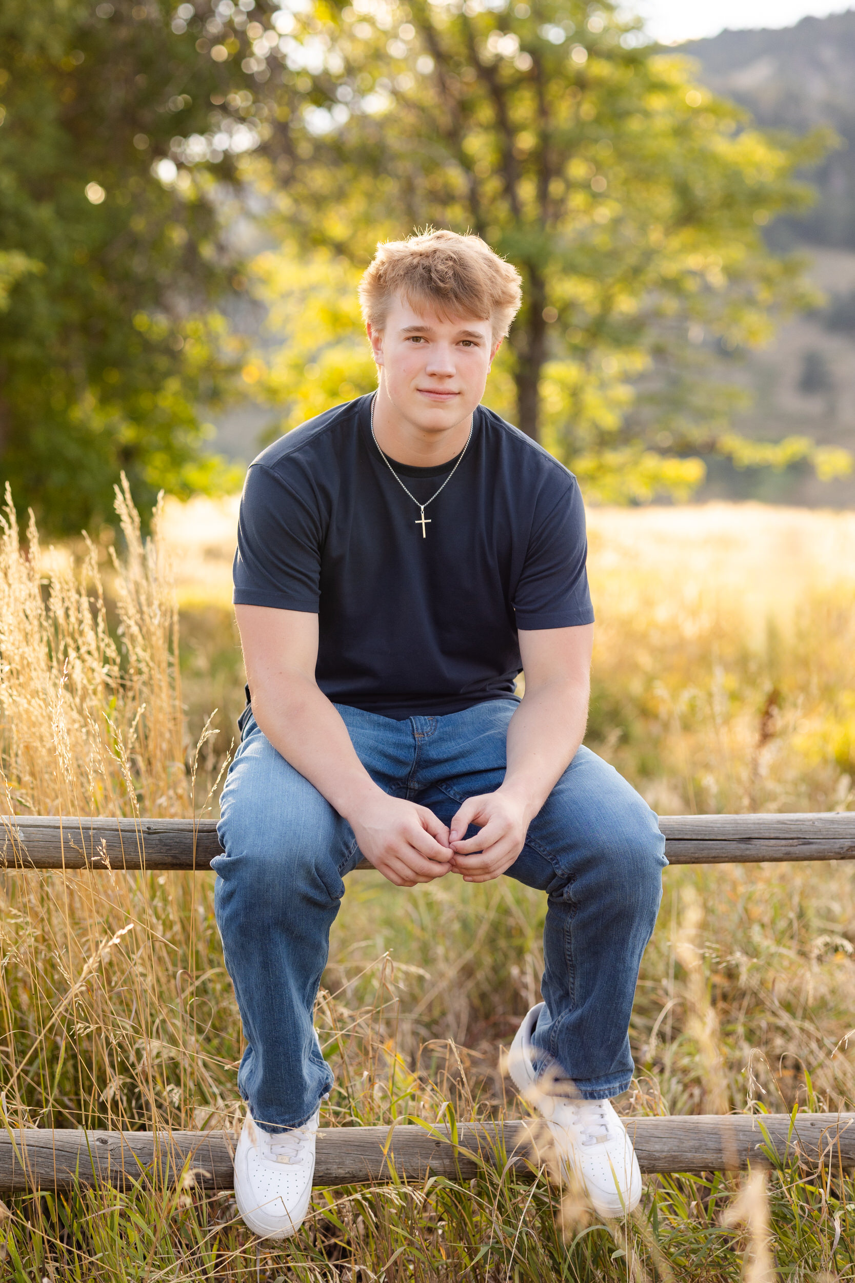 A young man sits on a fence at Chautauqua Park in Boulder, Colorado and smiles at the camera.
