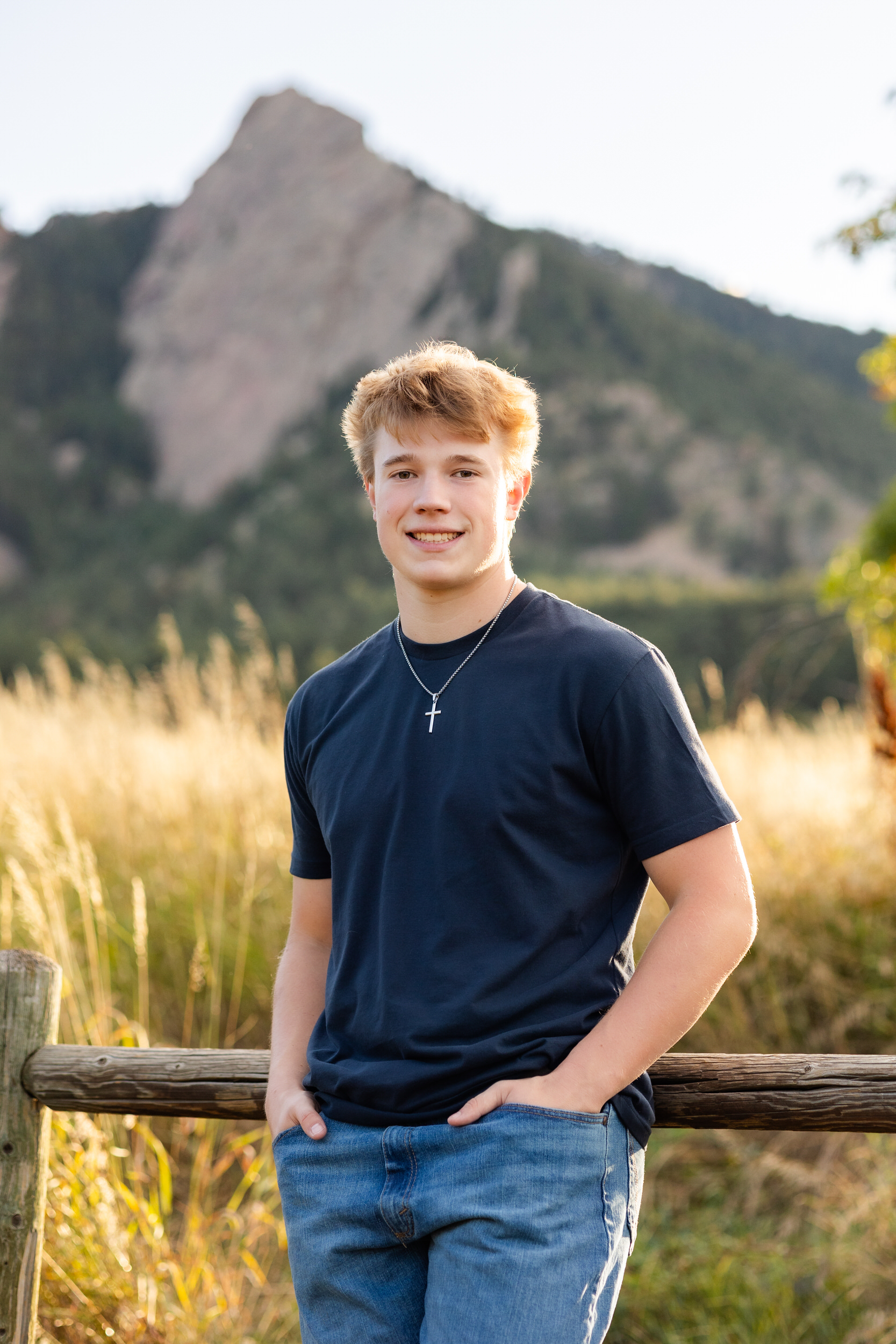 A young man leans against a fence in Chautauqua Park in Boulder, Colorado and smiles at the camera.