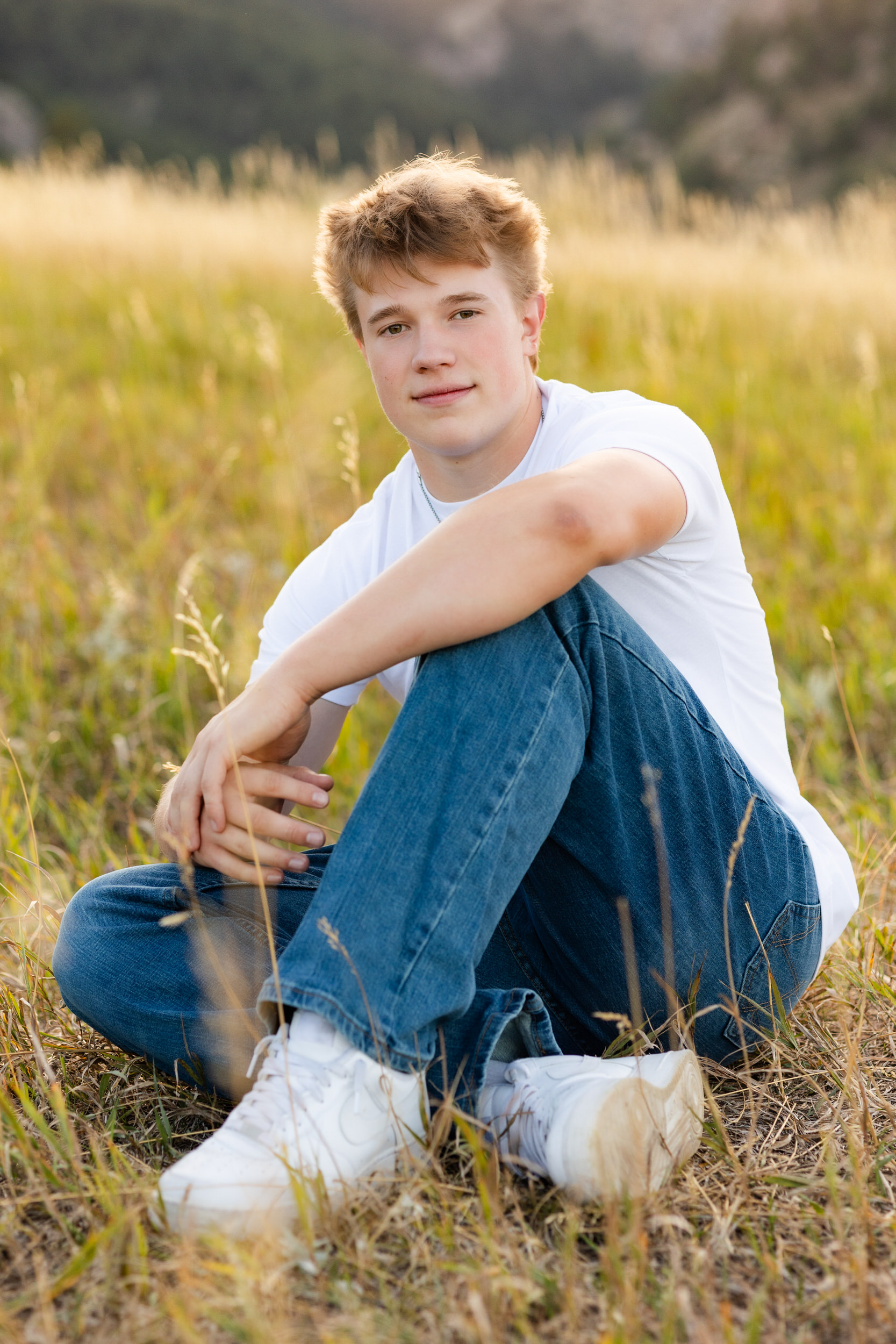 A young man sits in a field of tall grass with one arm resting on his knee and looks at the camera.