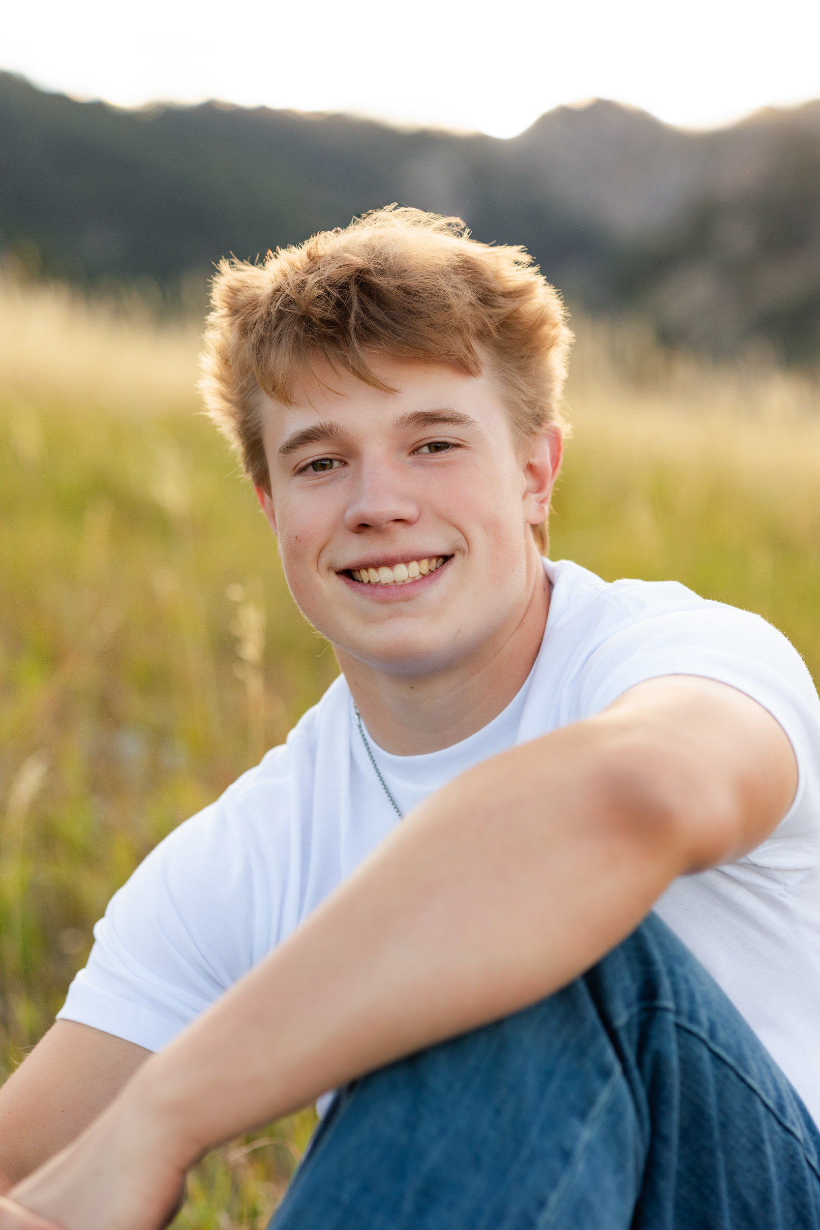 A young man sits in a field of tall grass with one arm resting on his knee and smiles at the camera.